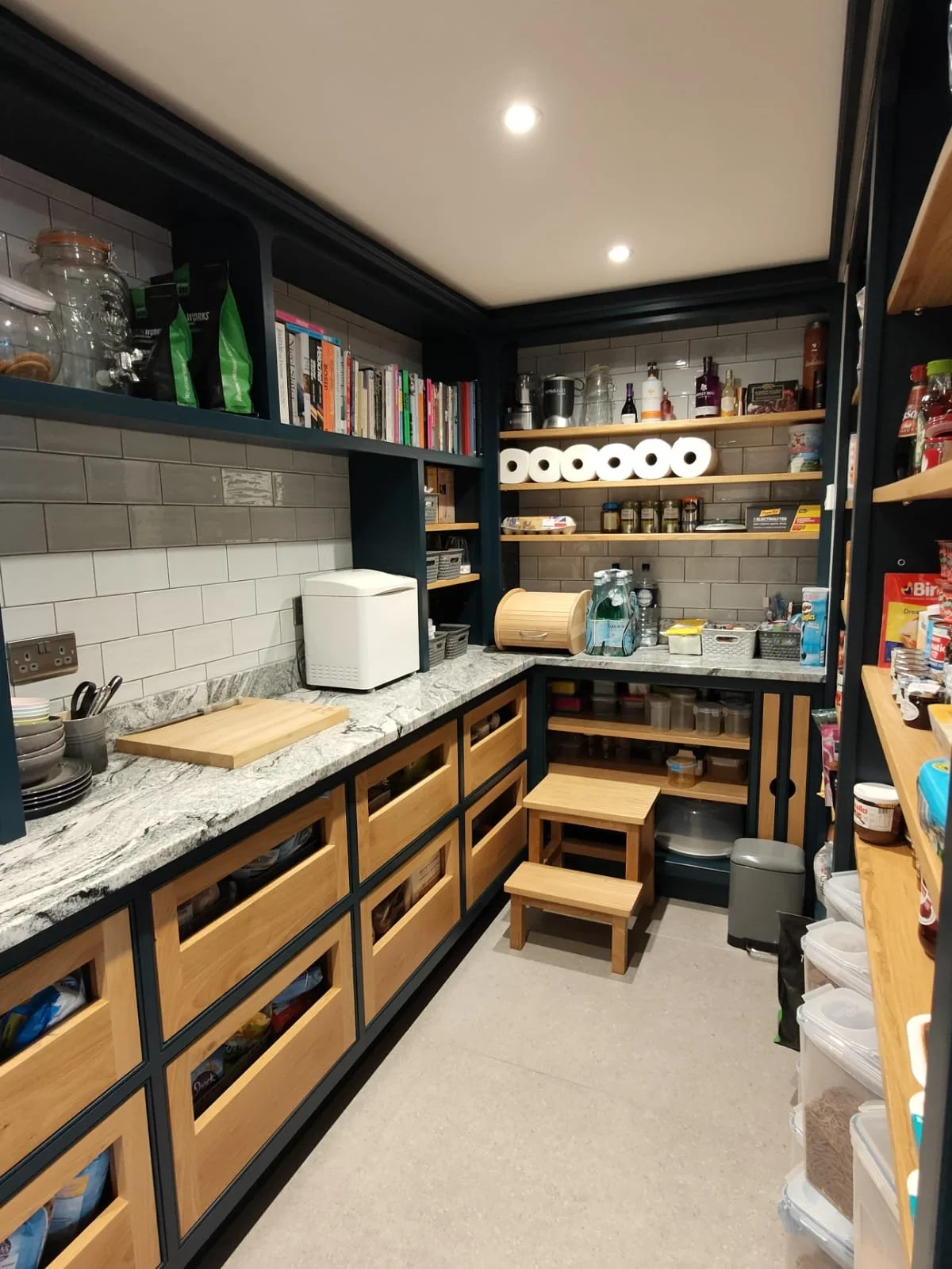 A well-organized pantry with wooden and black shelves, marble countertop, cutting board, food storage containers, books, and various pantry items, with gray tiled walls.