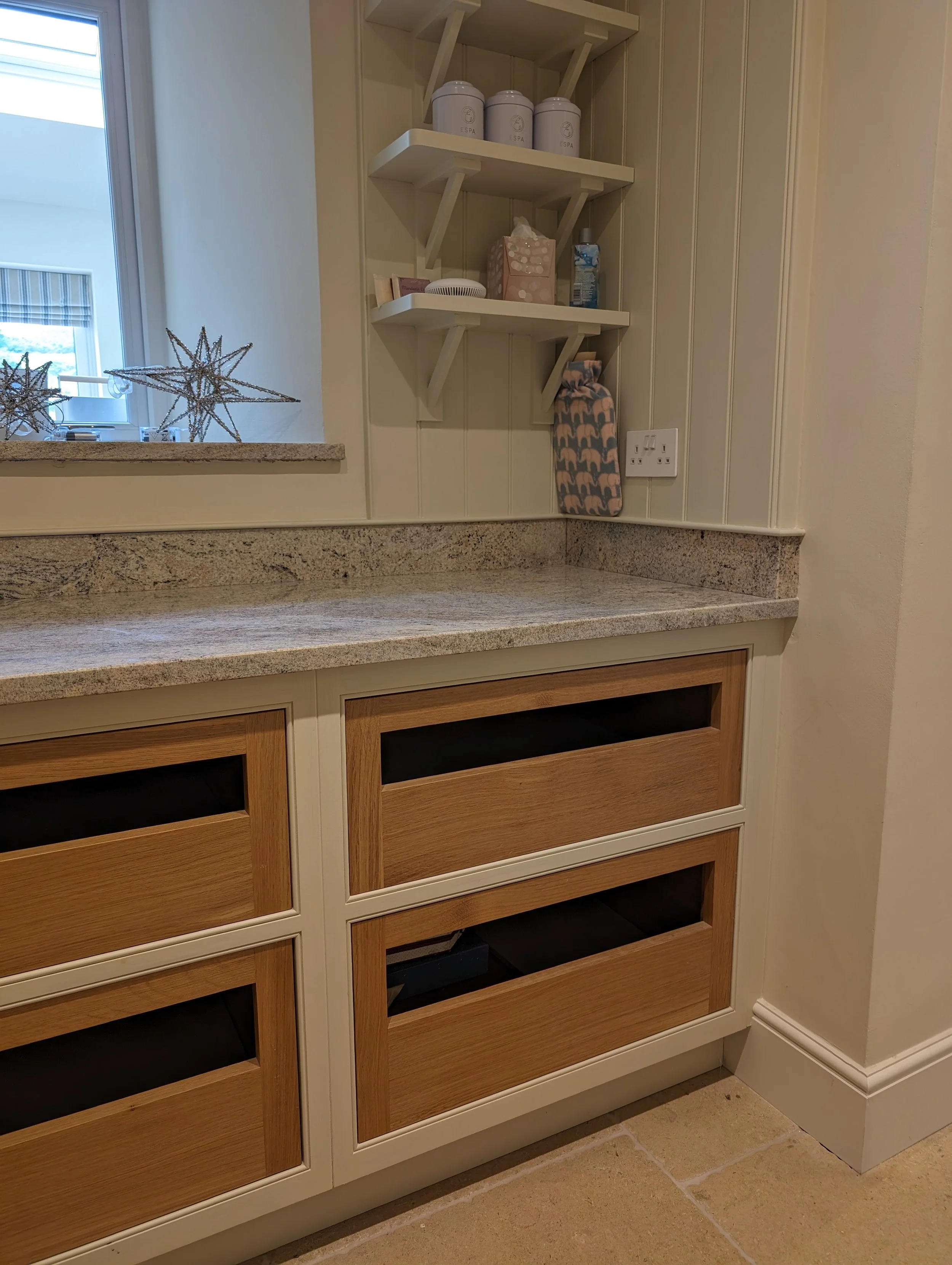 Kitchen countertop with wooden drawer units and wall shelves holding kitchen items, near a window with holiday star decorations.