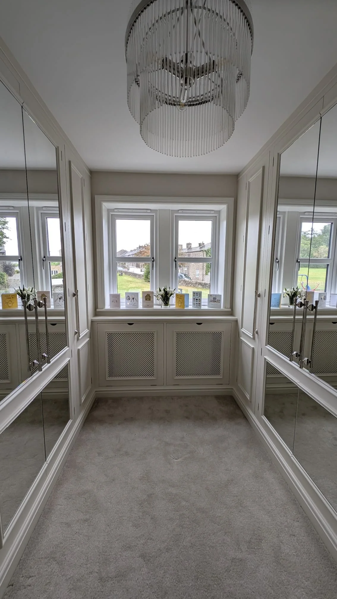 A bright, narrow room with white window frames and a ceiling chandelier. The windows reveal a green outdoor landscape. The room's walls have mirrors, and there is a ledge with decorative cards and small flower arrangements. The floor is covered with 