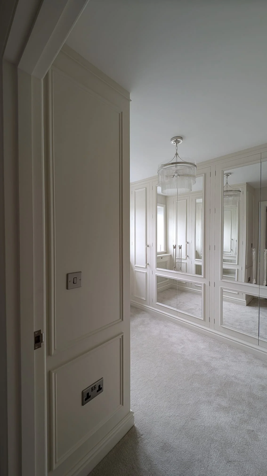 Luxury bedroom with white built-in wardrobes, mirrored doors, chandelier, and beige carpeted flooring.