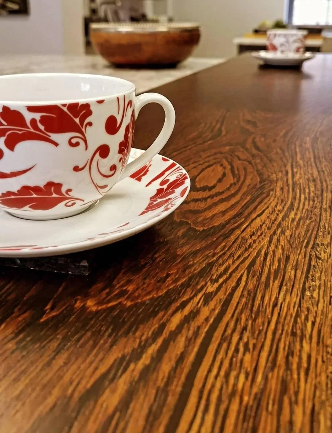 Close-up of a white teacup with red floral pattern and matching saucer on a wooden table. In the background, there are additional teacups and a wooden bowl.