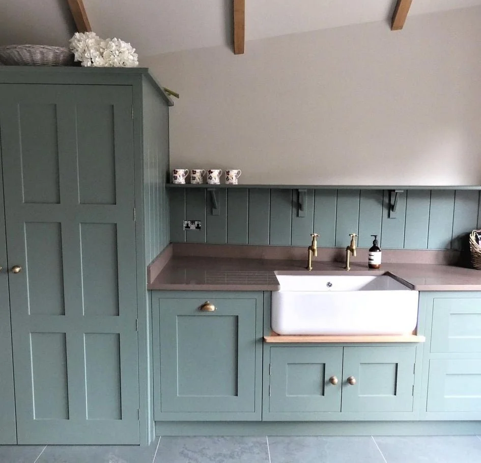 Kitchen with green cabinets, a farmhouse sink, four cups on a shelf, brass faucets, soap dispenser, and a laundry basket.