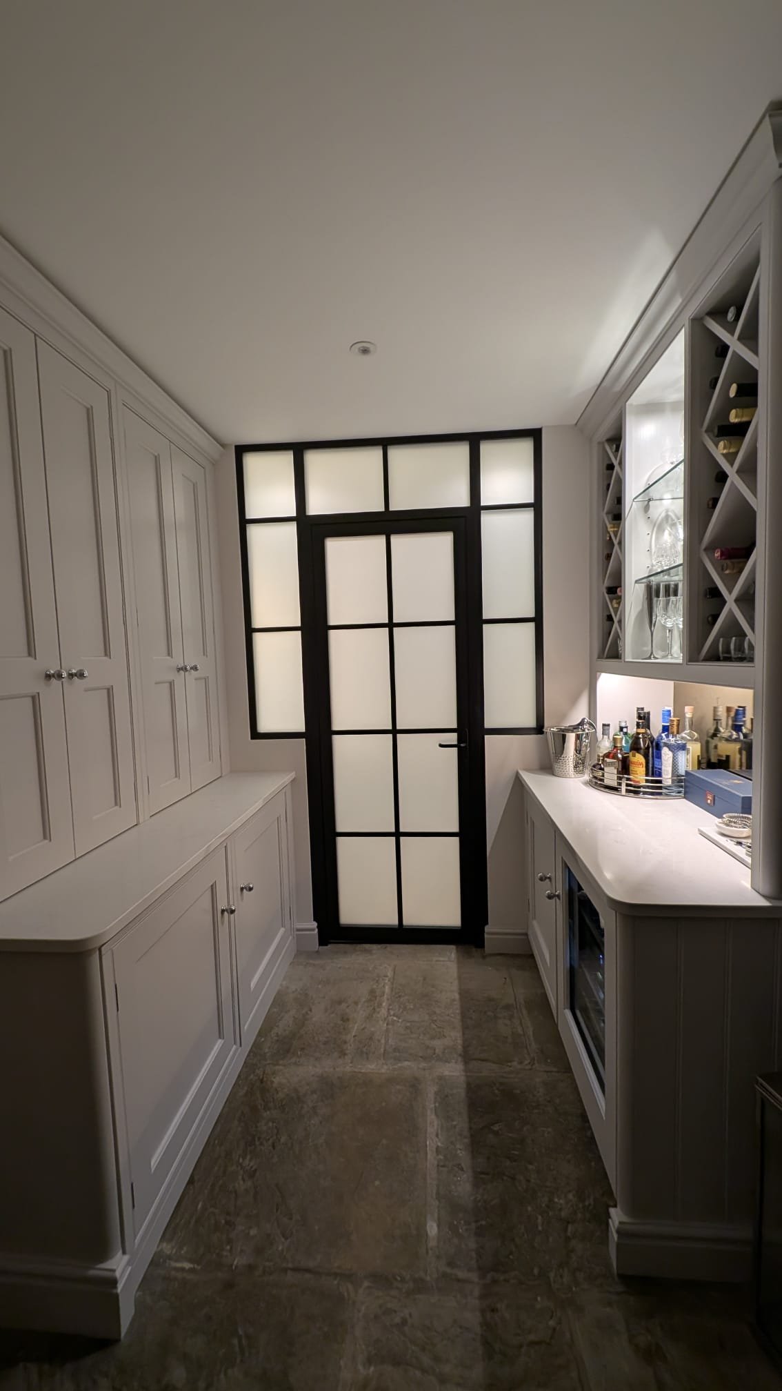 A modern bar area with white cabinetry on the sides, a black-framed glass door at the back, and a bar countertop with bottles and glasses.