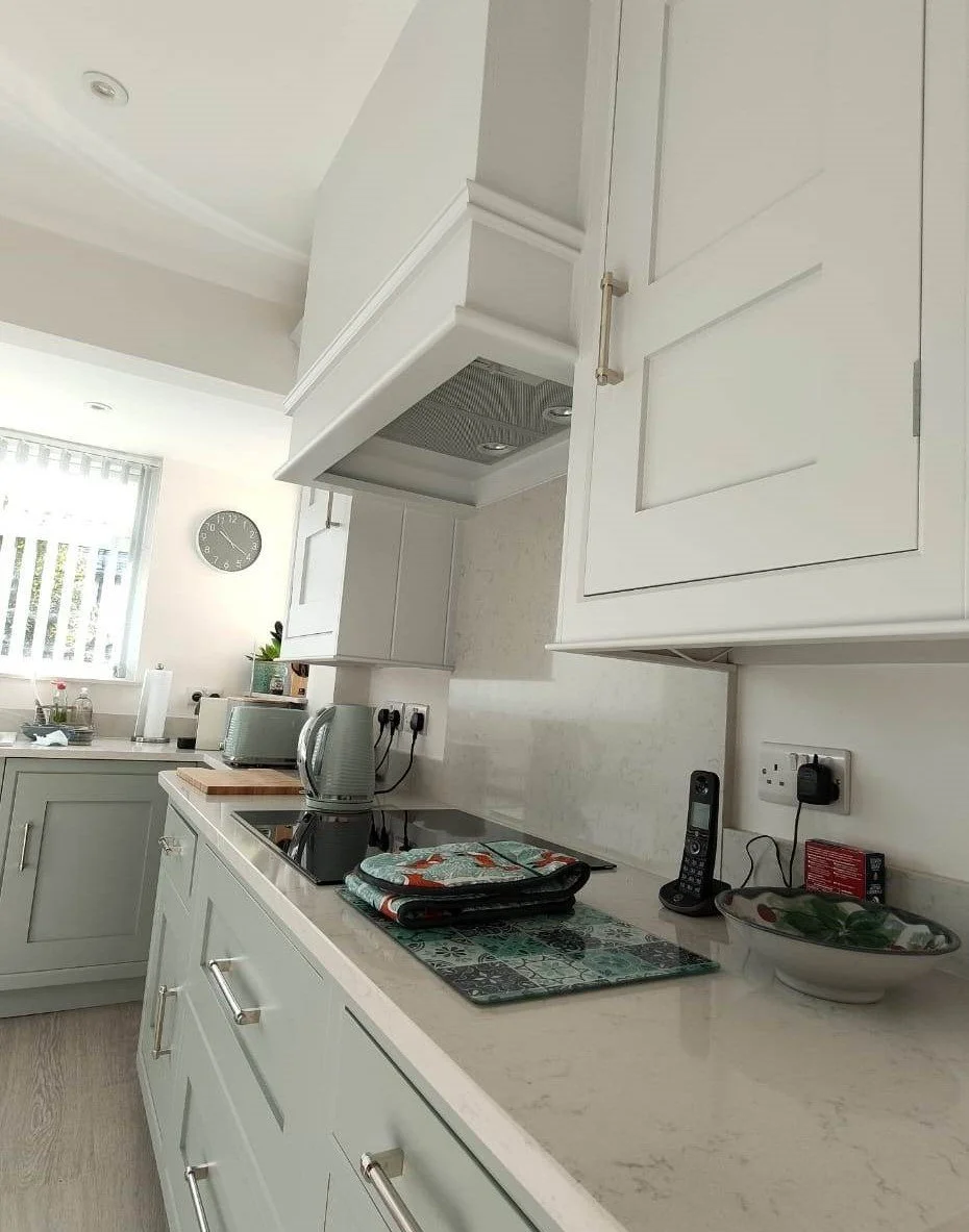 White kitchen with cabinets, countertop, and appliances including a kettle, toaster, and cordless phone. A bowl with salad is on the counter, with a clock on the wall and a window with vertical blinds.