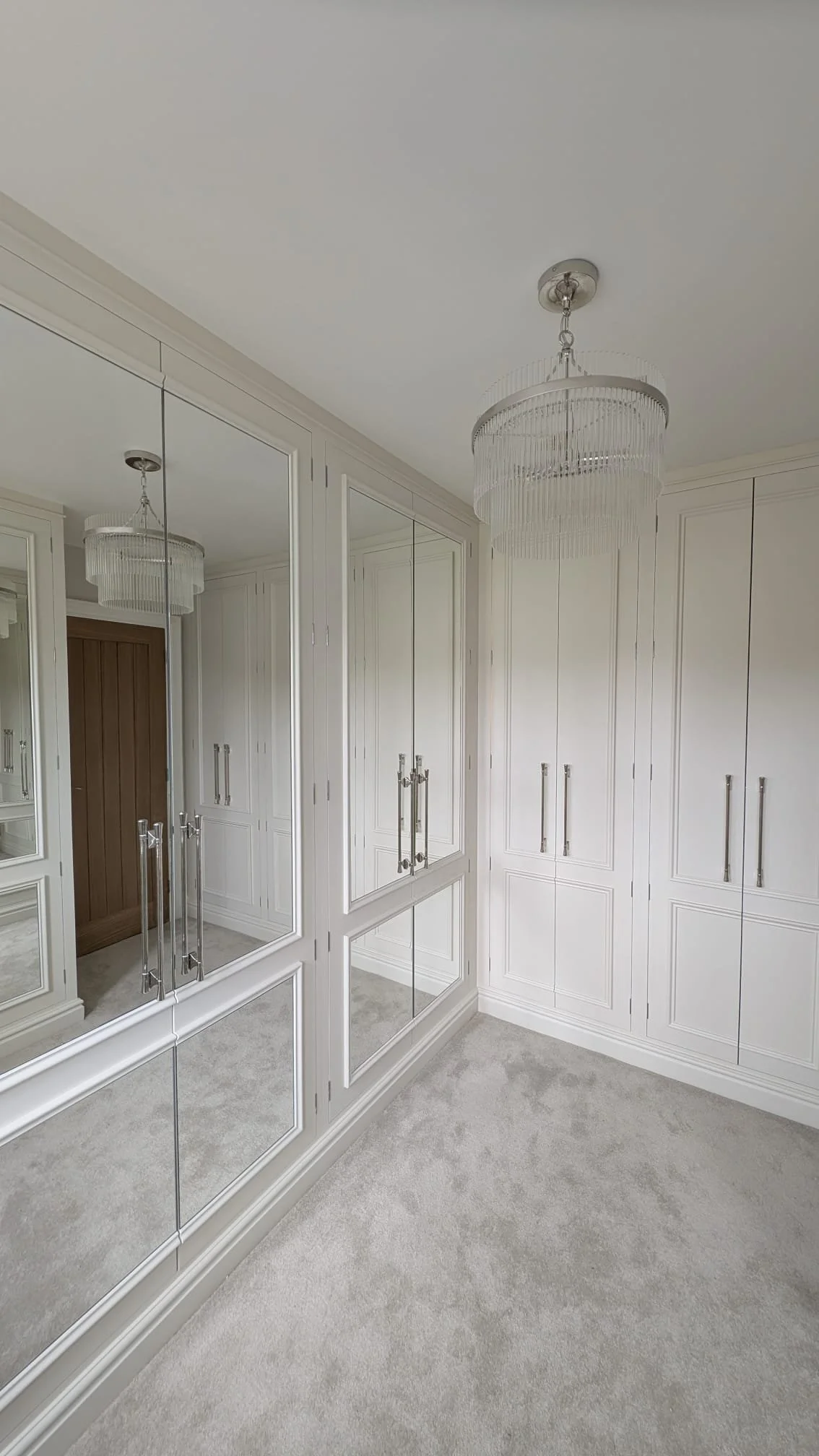A walk-in closet with white cabinetry and mirrored sliding doors, a white ceiling, beige carpet, and a chandelier with hanging crystals.