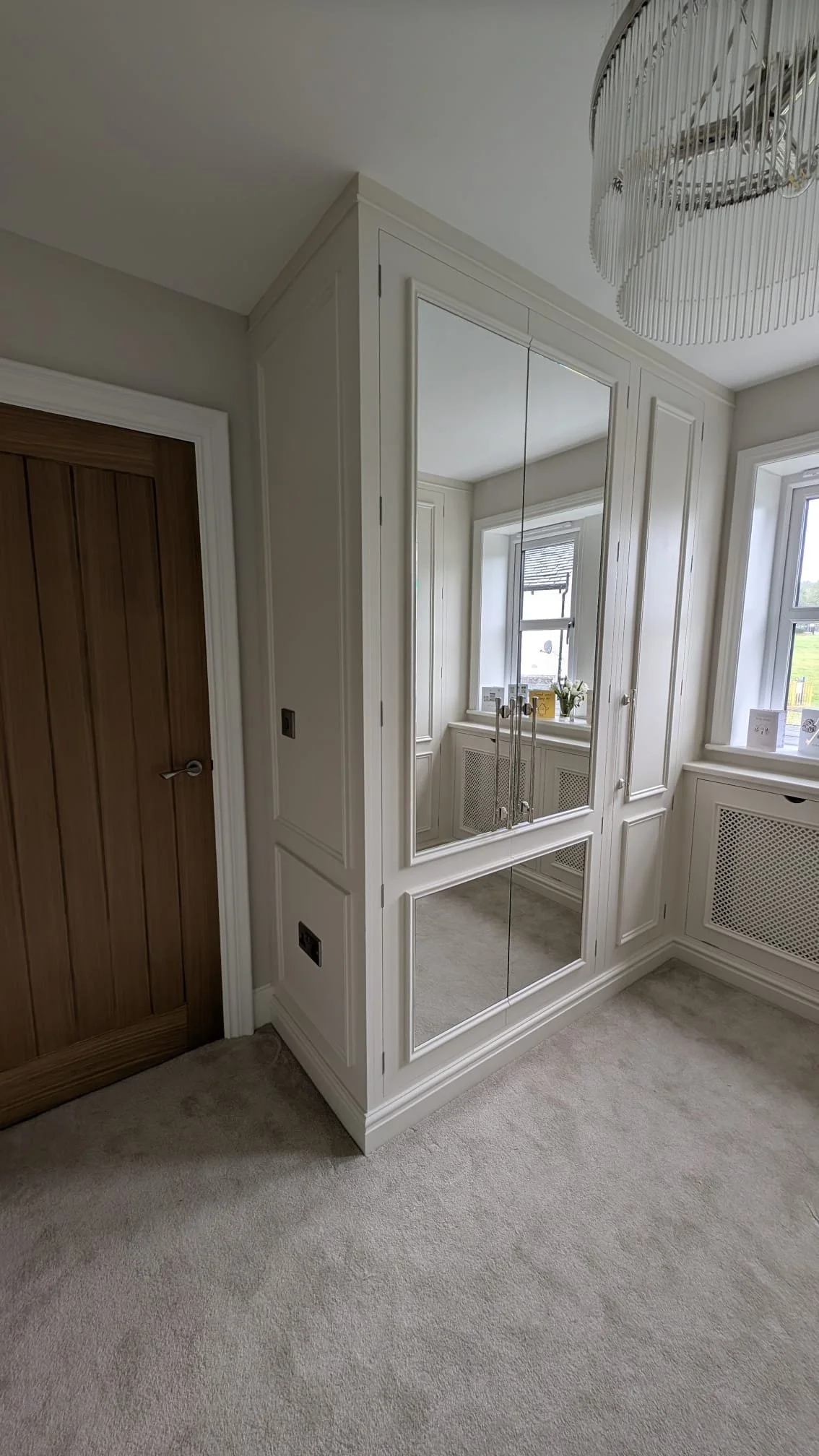 A corner of a room with beige carpet, a white paneled wall with built-in cabinetry, mirrored doors, and a window with a radiator cover beneath it.