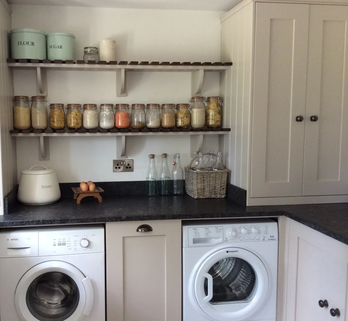 A laundry room with a black countertop, white cabinets, a washing machine, and shelves holding jars of pasta, sugar, flour, and eggs.