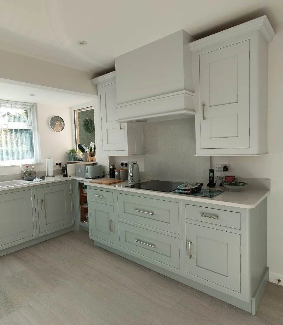 Kitchen with light green cabinets, a window, a clock, appliances including a microwave, toaster, kettle, and electric stove, and various small items on the countertops.