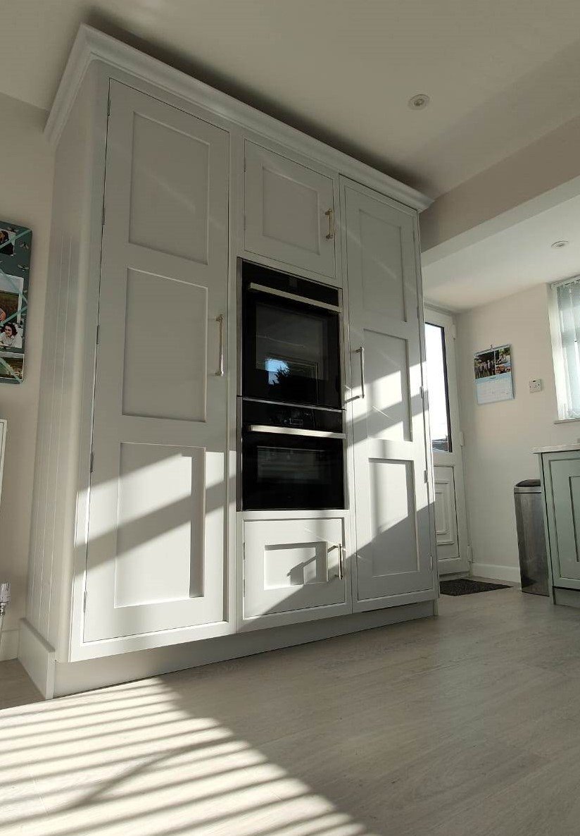 A kitchen with white cabinets and built-in black double oven, sunlight casting shadows on the floor.