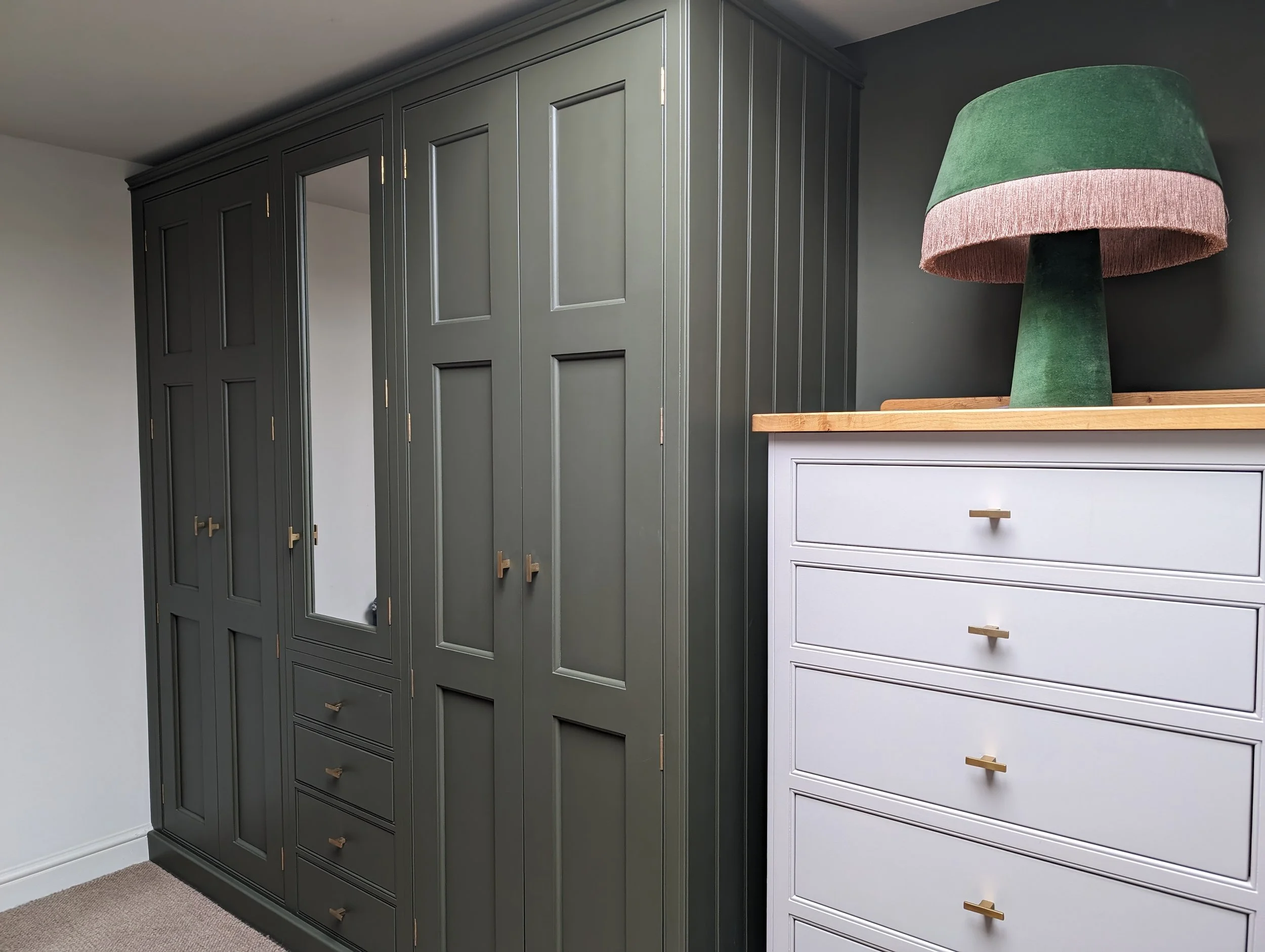 A bedroom with a dark green wardrobe, a white chest of drawers with gold handles, and a green and pink lampshade on top of the chest of drawers.