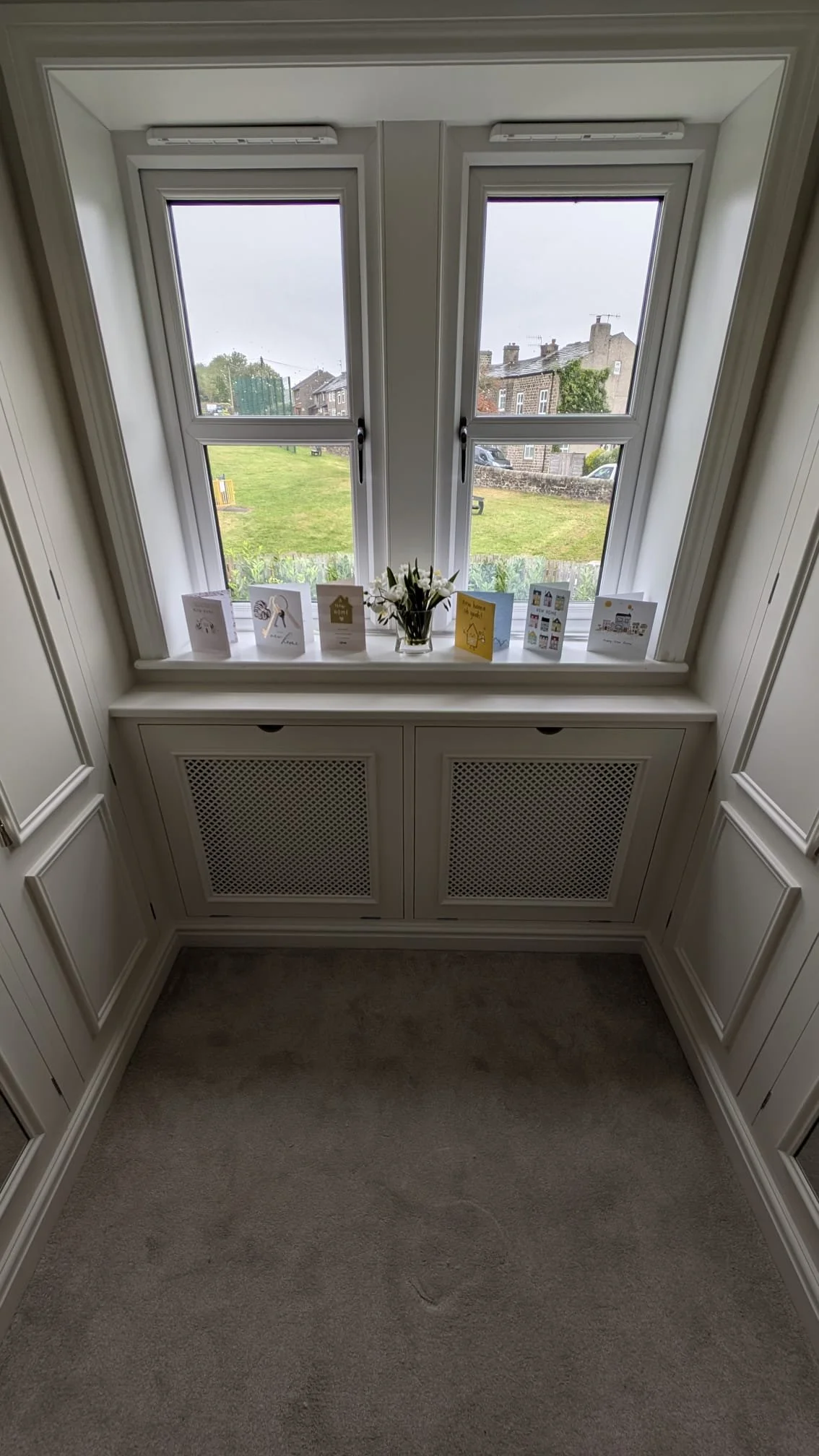 View through a window showing a grassy yard with houses in the background, decorated with greeting cards and a small vase of flowers on the windowsill.