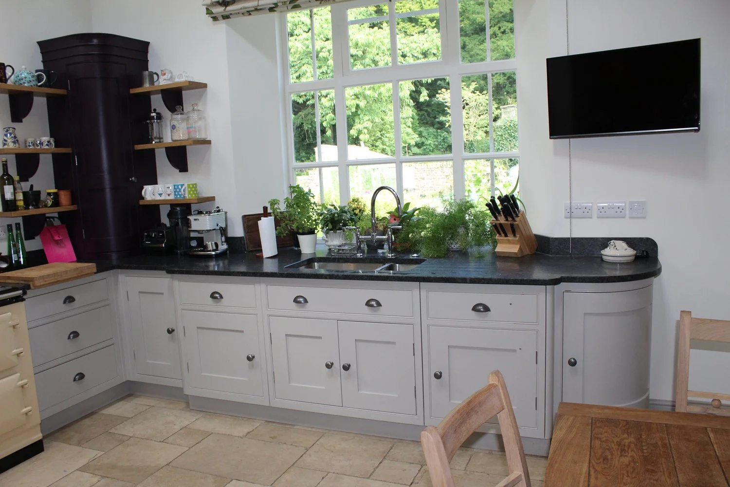Kitchen with white cabinets, black granite countertop, window above sink with green trees outside, open shelves with cups and jars, coffee machine, cutting board, knife block, potted plants by the window, TV on wall, chairs, and tiled floor.