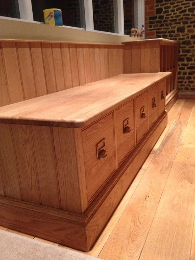 Wooden bench with six drawers underneath, placed against a wooden-paneled wall in a room with hardwood flooring and brick wall in the background.
