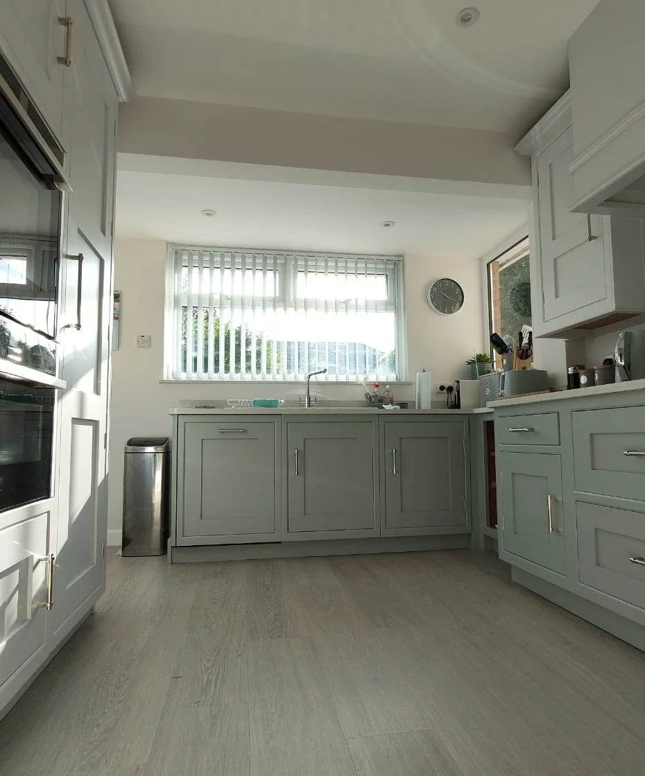 Clean, modern kitchen with light gray cabinets, a white countertop, and a window with vertical blinds, outside view of greenery, clock on wall, and various kitchen appliances and utensils.