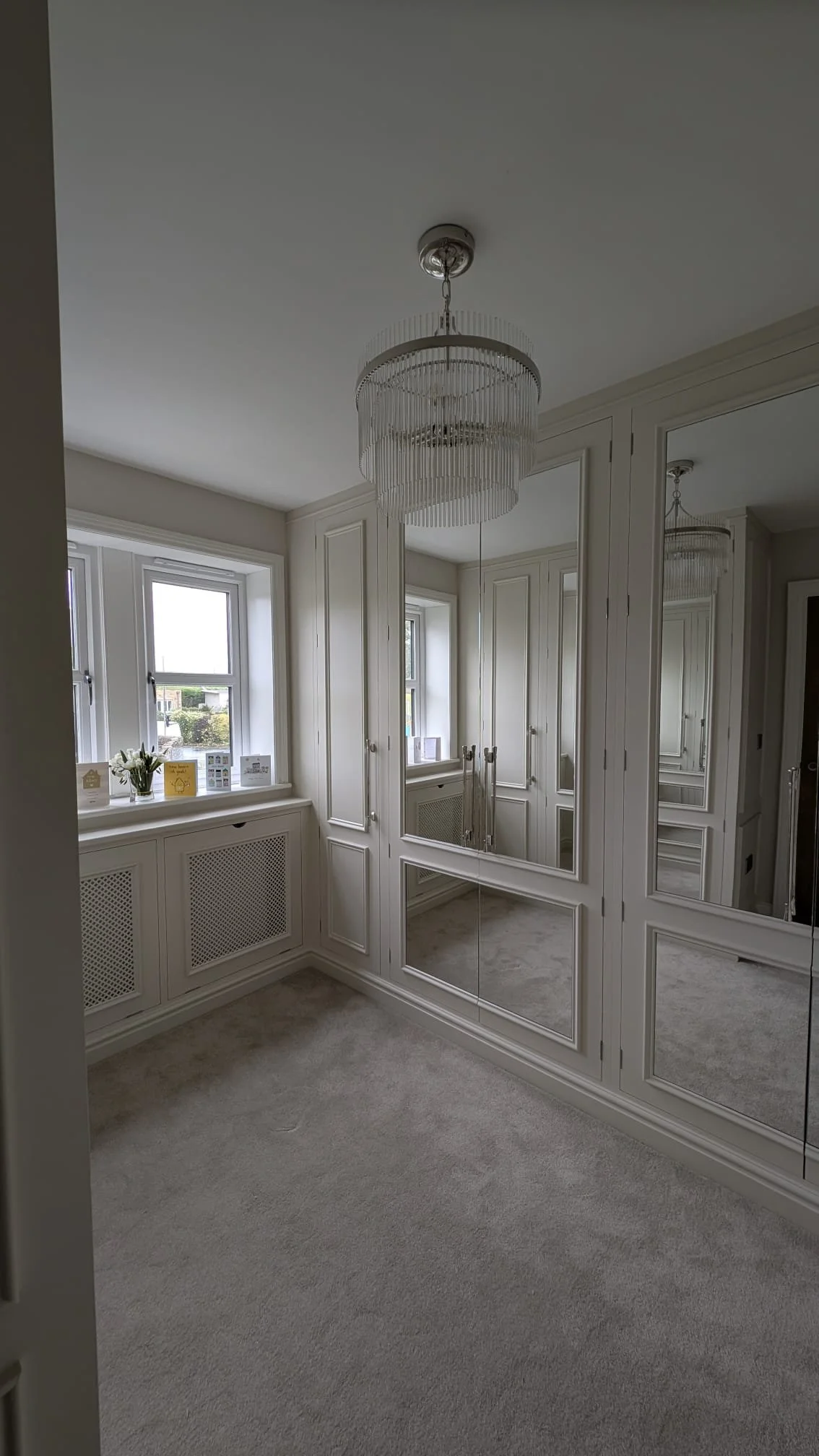 A corner of a room with white cabinetry, mirrored closet doors, a window with a vase of flowers and greeting cards on the window sill, light-colored carpet, and a white chandelier hanging from the ceiling.