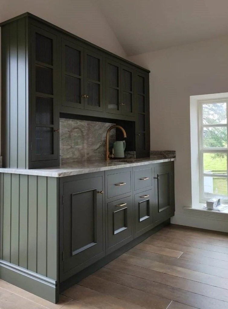 Kitchen with dark green cabinets, marble countertop, gray marble backsplash, brass faucet, and a window with a view of green trees outside.