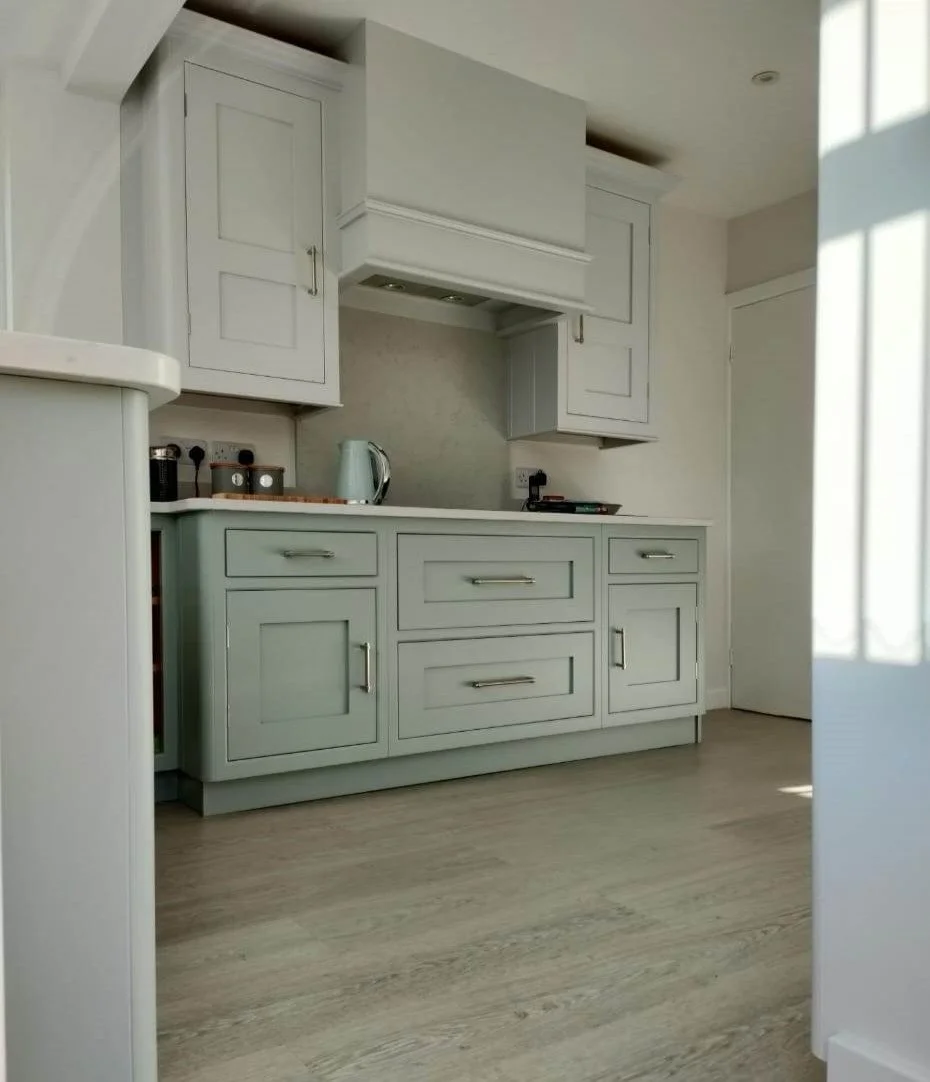 A kitchen with white cabinets, a countertop, and a wooden floor. There is a kettle and some small containers on the countertop. Part of a doorway is visible on the right, with sunlight coming through a window.