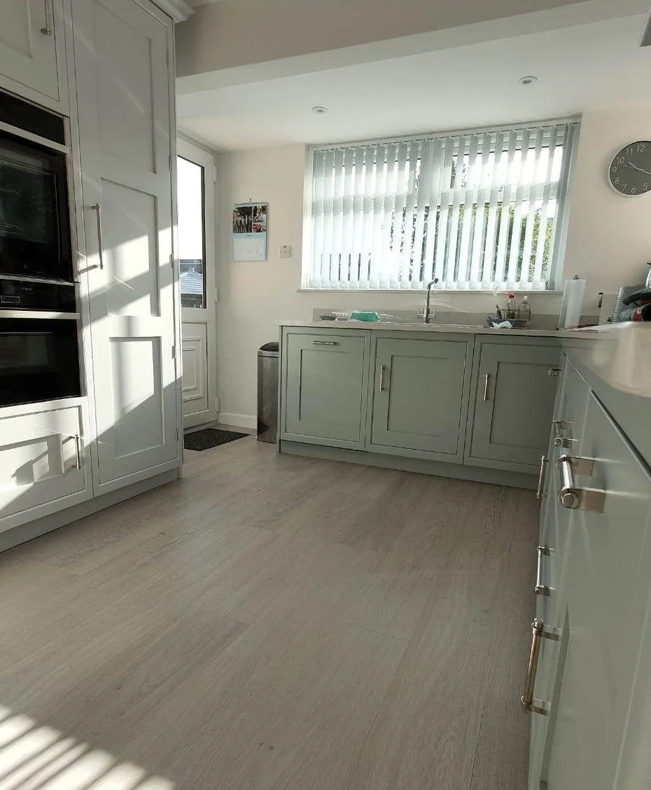Bright kitchen with white cabinets, a large window with vertical blinds, a clock on the wall, and a countertop with small appliances and dishes.