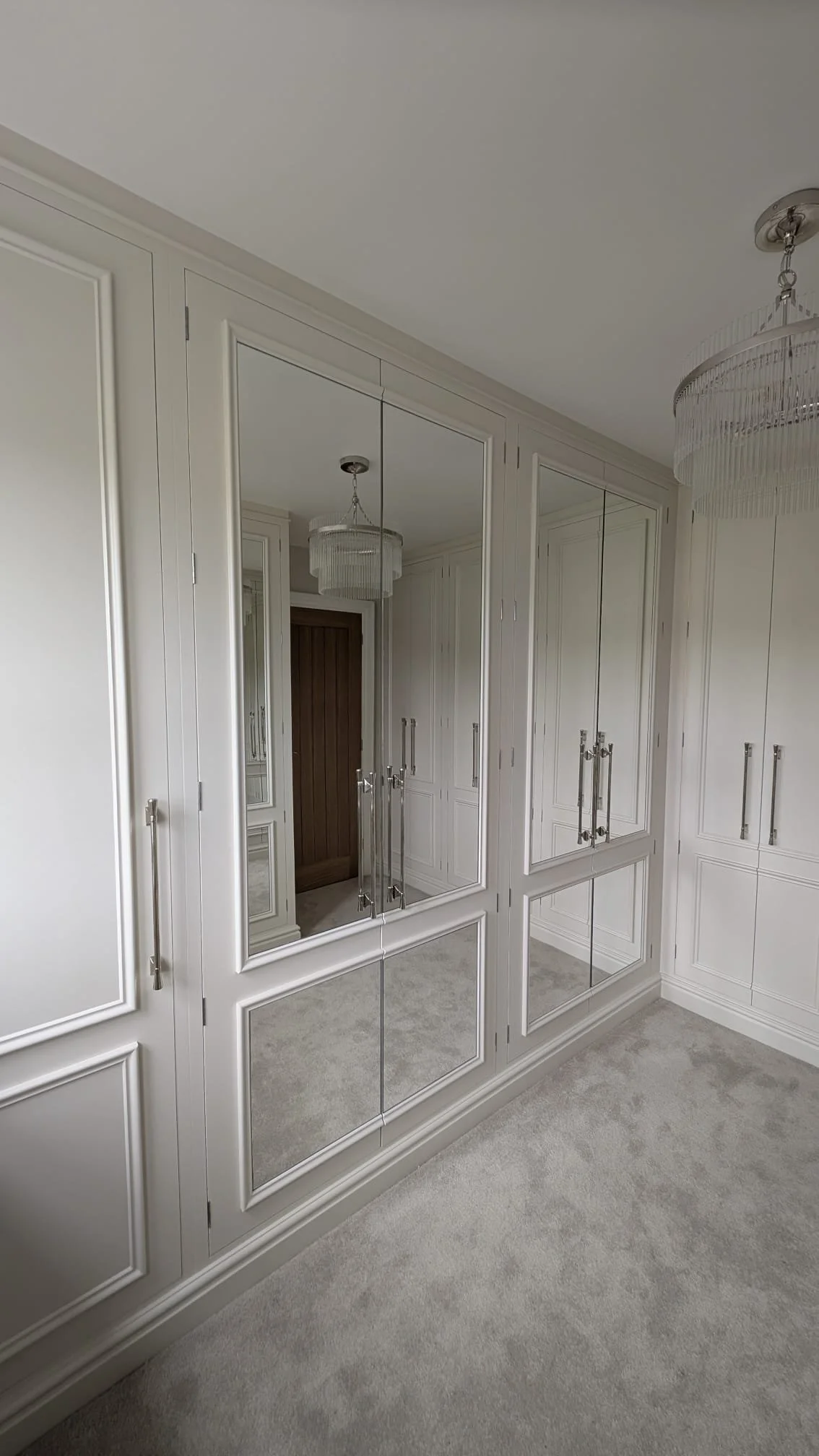 Interior view of a closet with white built-in cabinets and large mirrored doors, carpeted floor, and a chandelier ceiling light.