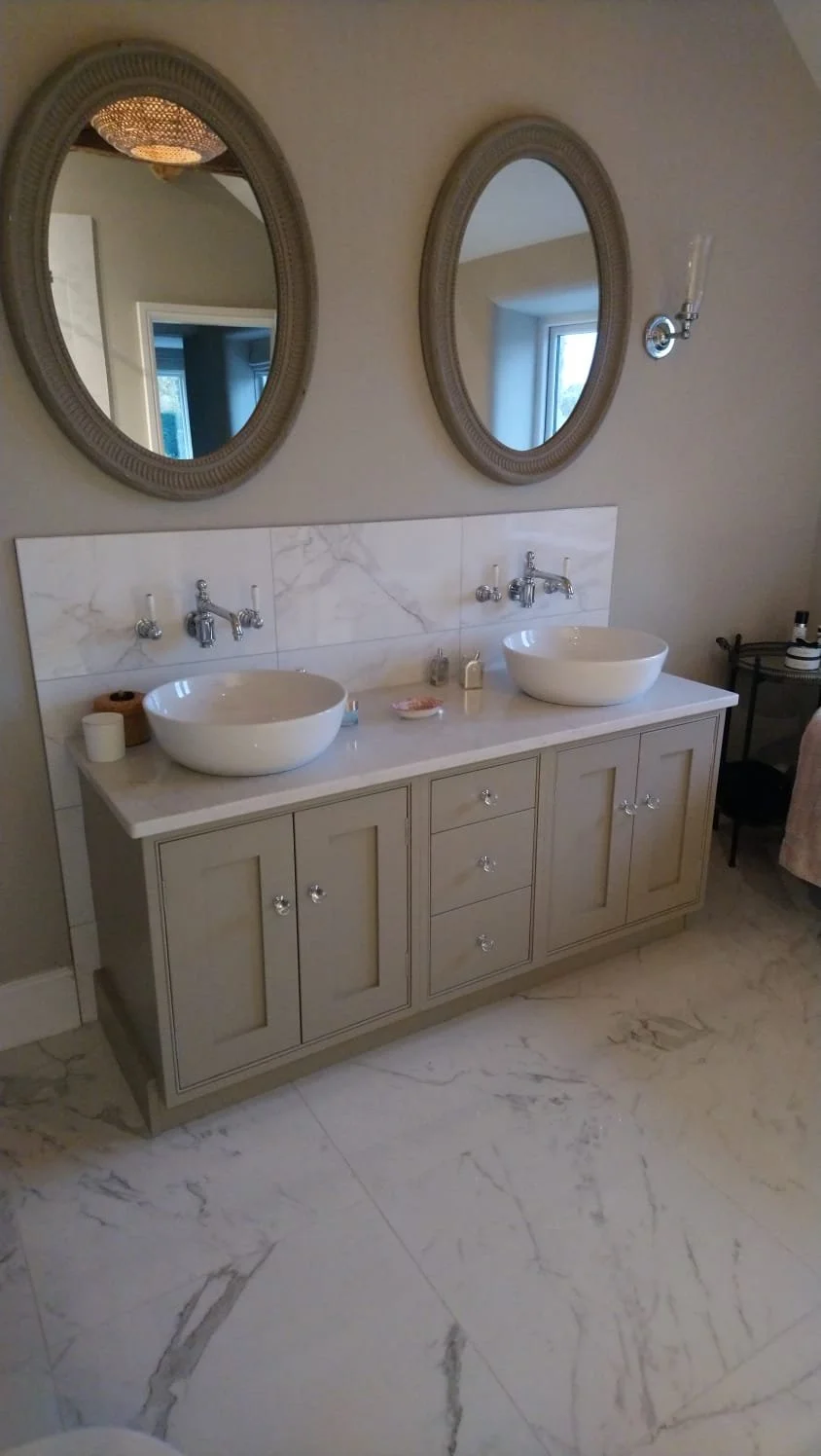 A bathroom with dual oval mirrors above a beige vanity with two vessel sinks and marble countertop, with wall-mounted faucets and white marble tile backsplash.