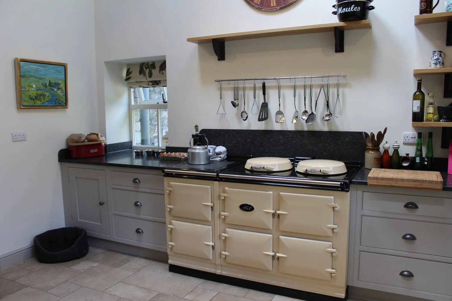 A vintage cream-colored AGA stove in a kitchen with black countertops, gray cabinets, hanging kitchen utensils, a window, and various kitchen items on shelves and counters.