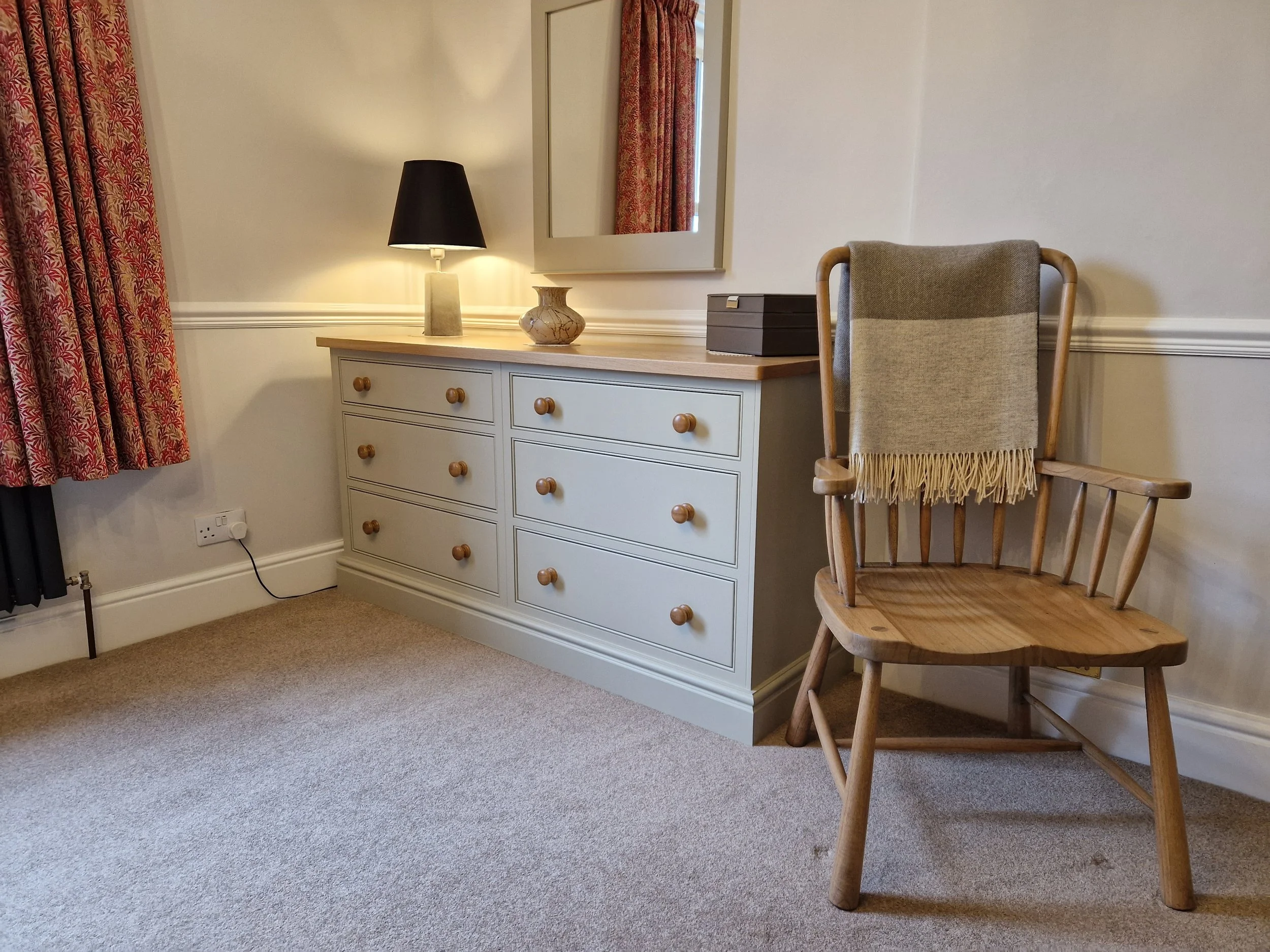 A bedroom corner with a white dresser topped with a black lamp, a small decorative vase, and a black box. A mirror hangs above the dresser. To the right, there is a wooden chair with a beige and grey fringed throw blanket draped over the back.