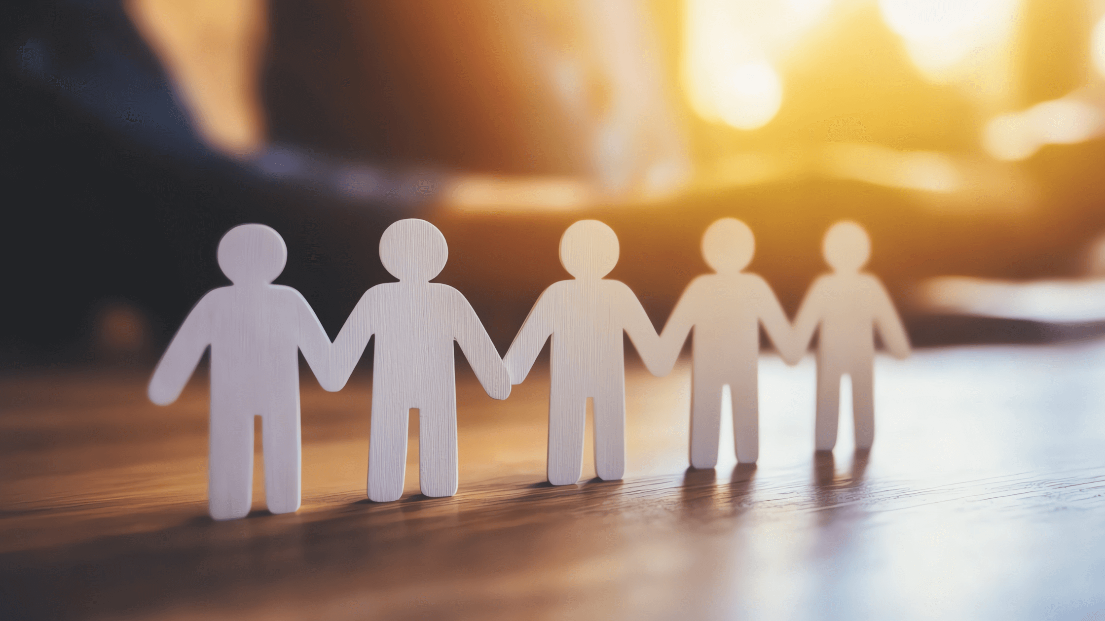 Figures of a group of people lined up, holding hands on a wooden table at sunset.