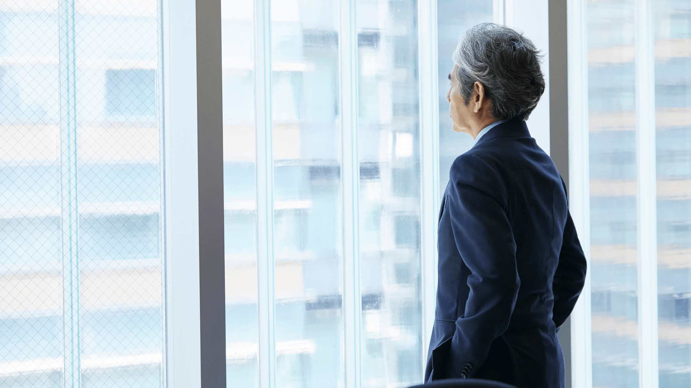 A person looking out of a city window, in a navy blue suit during the day.