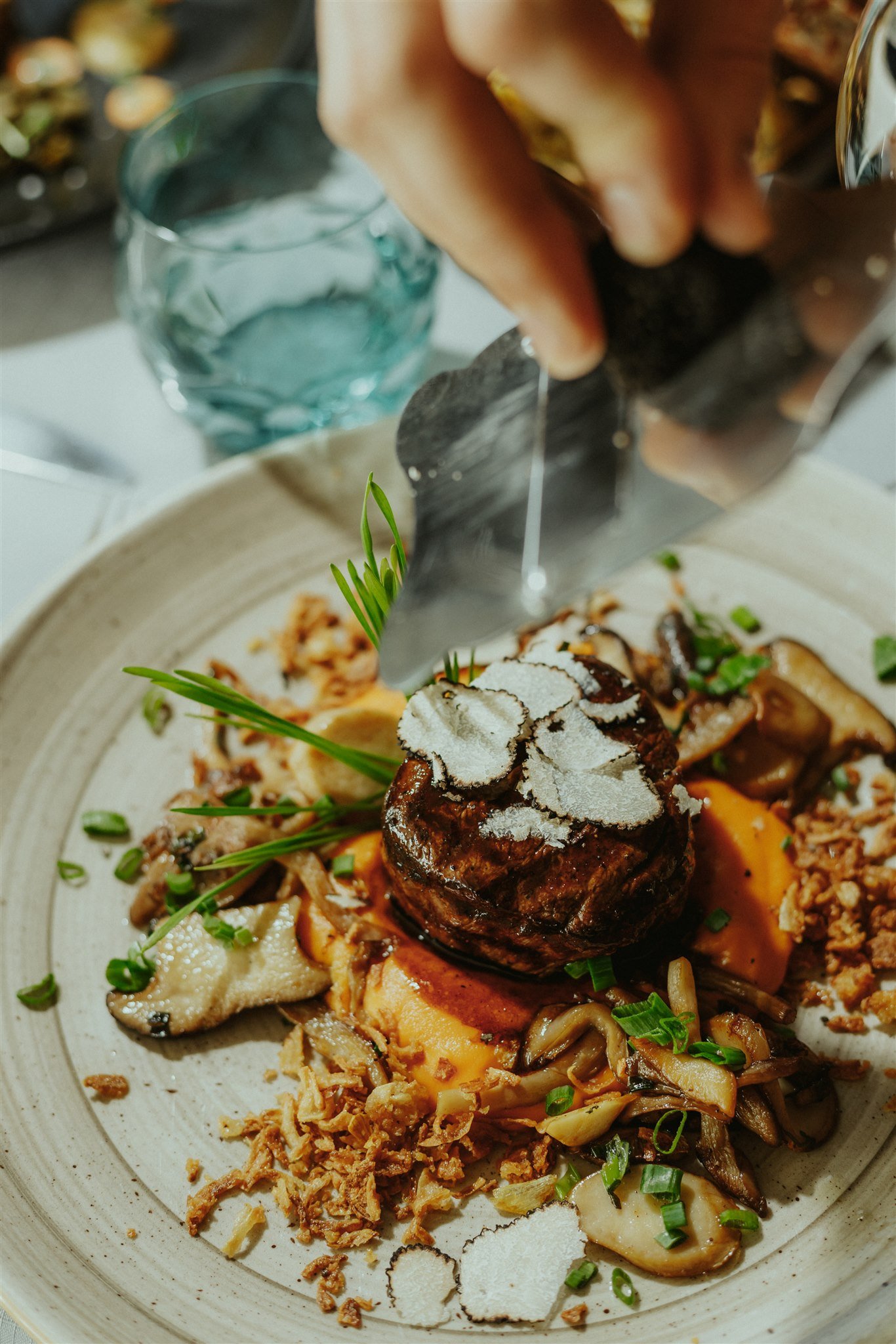 A gourmet dish featuring a beef steak topped with shaved truffles, surrounded by garnishes of mushrooms, crispy onions, and green herbs on a rustic plate. A hand is visible grating truffles over the steak.