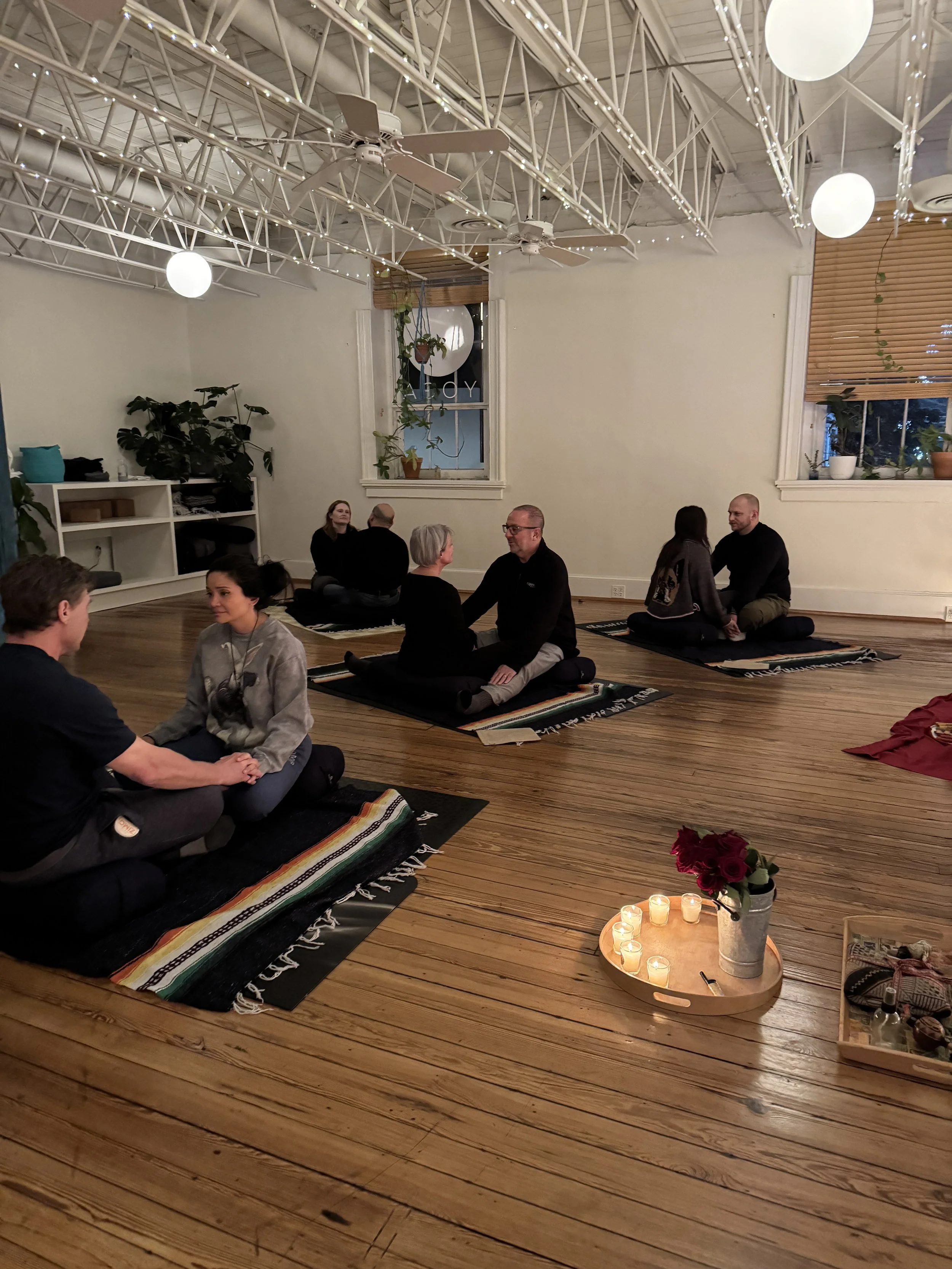 Several couples participate in a communication activity while seated on cushions in a cozy room with soft lighting and yoga mats.