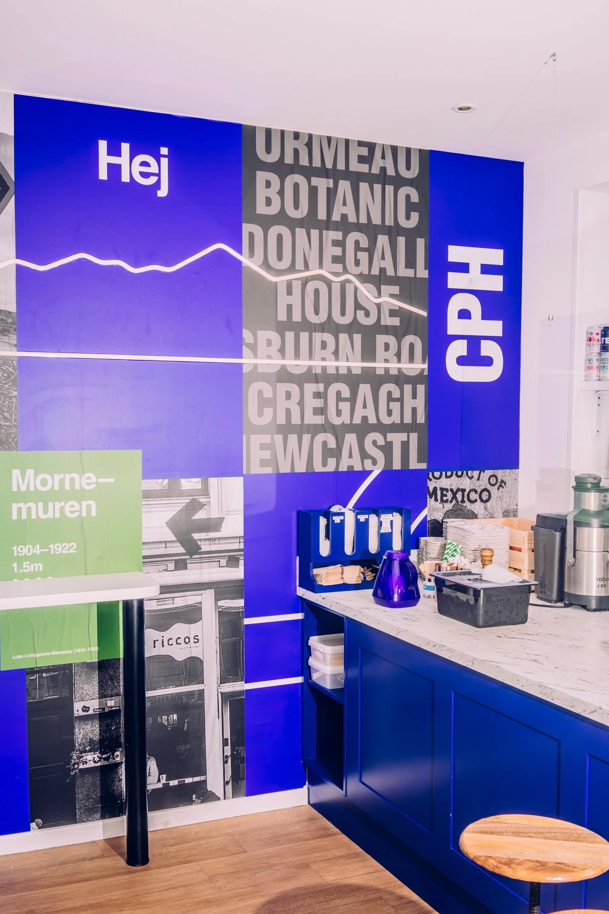 A brightly colored cafe or snack bar with a large blue wall featuring white and gray text and graphics, a counter with various items including a purple pitcher, papers, a black container, a coffee machine, and a small wooden stool.