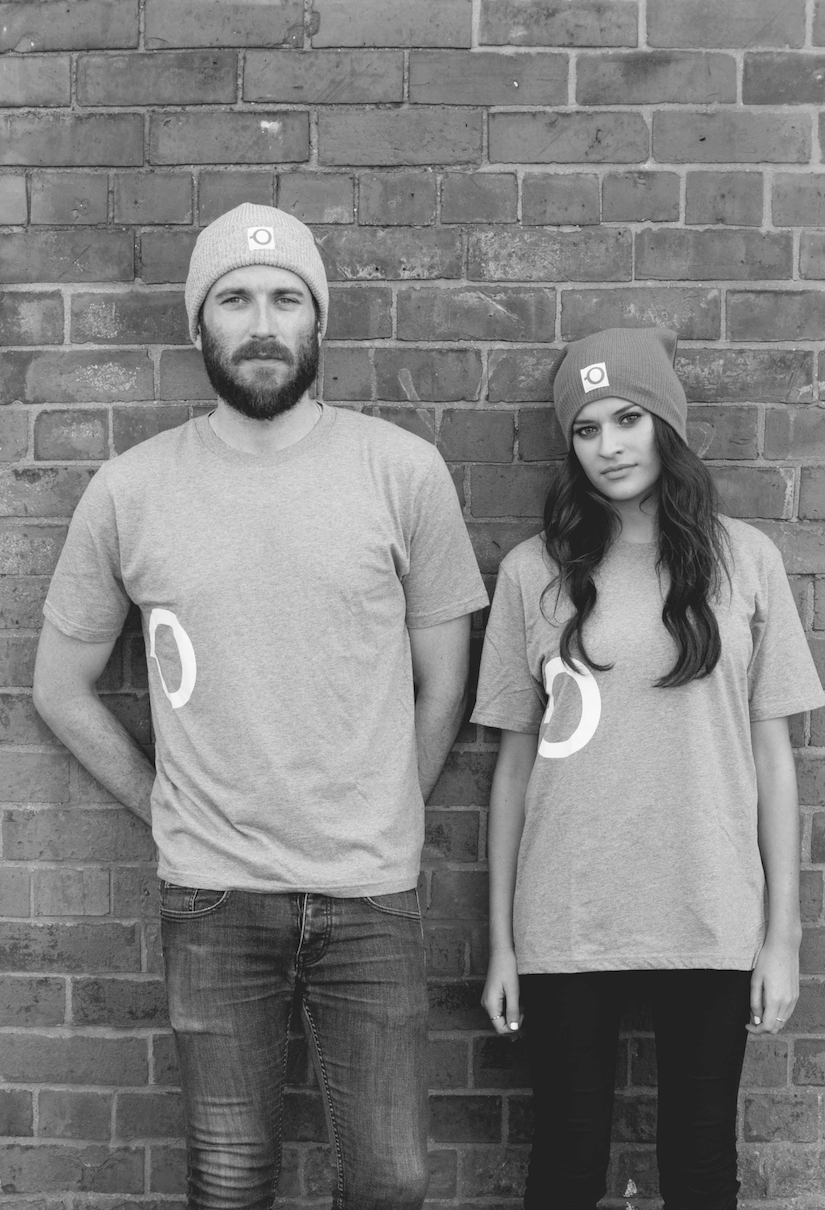 Two people, a man and a woman, standing against a brick wall. They are wearing beanies and casual t-shirts, and they are looking at the camera.