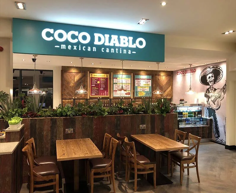Interior of a Mexican restaurant called Coco Diablo with wooden tables, chairs, greenery, and a wall with a drawing of a skeleton playing guitar.