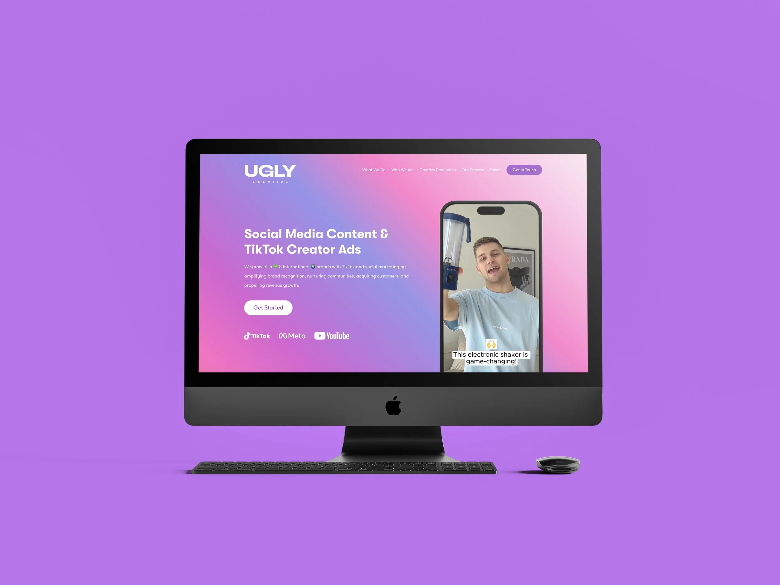 An Apple iMac with a keyboard and mouse in front, displaying a website for a creative agency called Ugly. The website promotes social media content and TikTok creator ads, with a video of a man holding an electronic shaker on a phone screen.