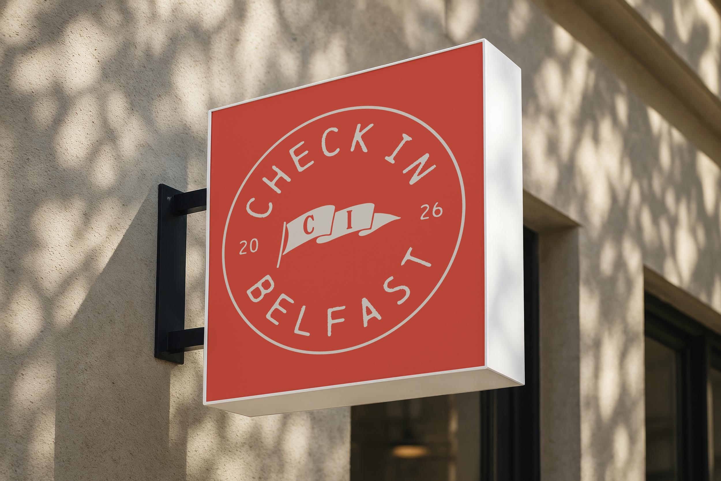 Red and white sign with a circular logo and text, mounted on a beige wall. Shadows of tree leaves are visible on the wall.