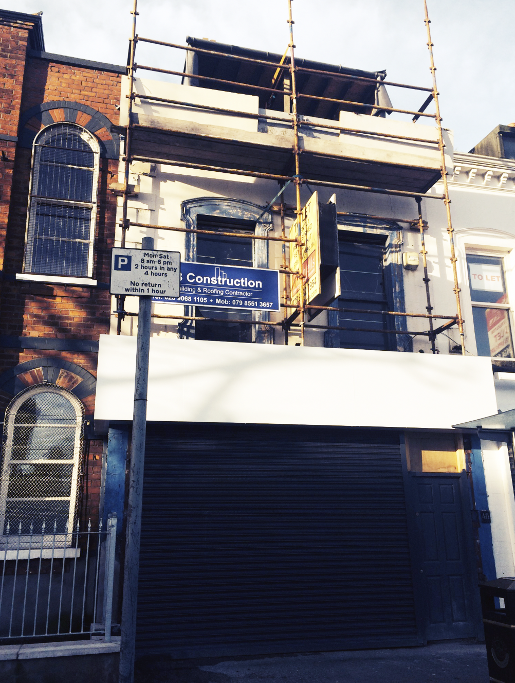 Building under construction with scaffolding, a construction sign, a roller shutter, and neighboring buildings with arched windows.