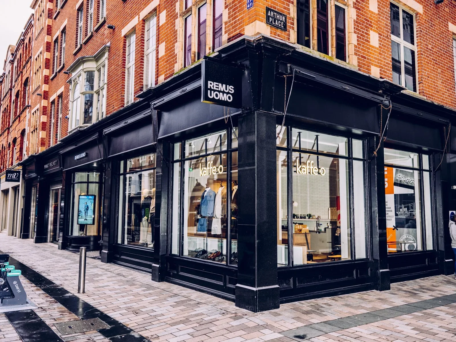 Street view of a clothing store with large glass windows, a black exterior, and mannequins wearing suits inside. The store is at the corner of a brick building on Arthur Place, with signs for "REMUS UOMO" and "kaffe-o" visible.