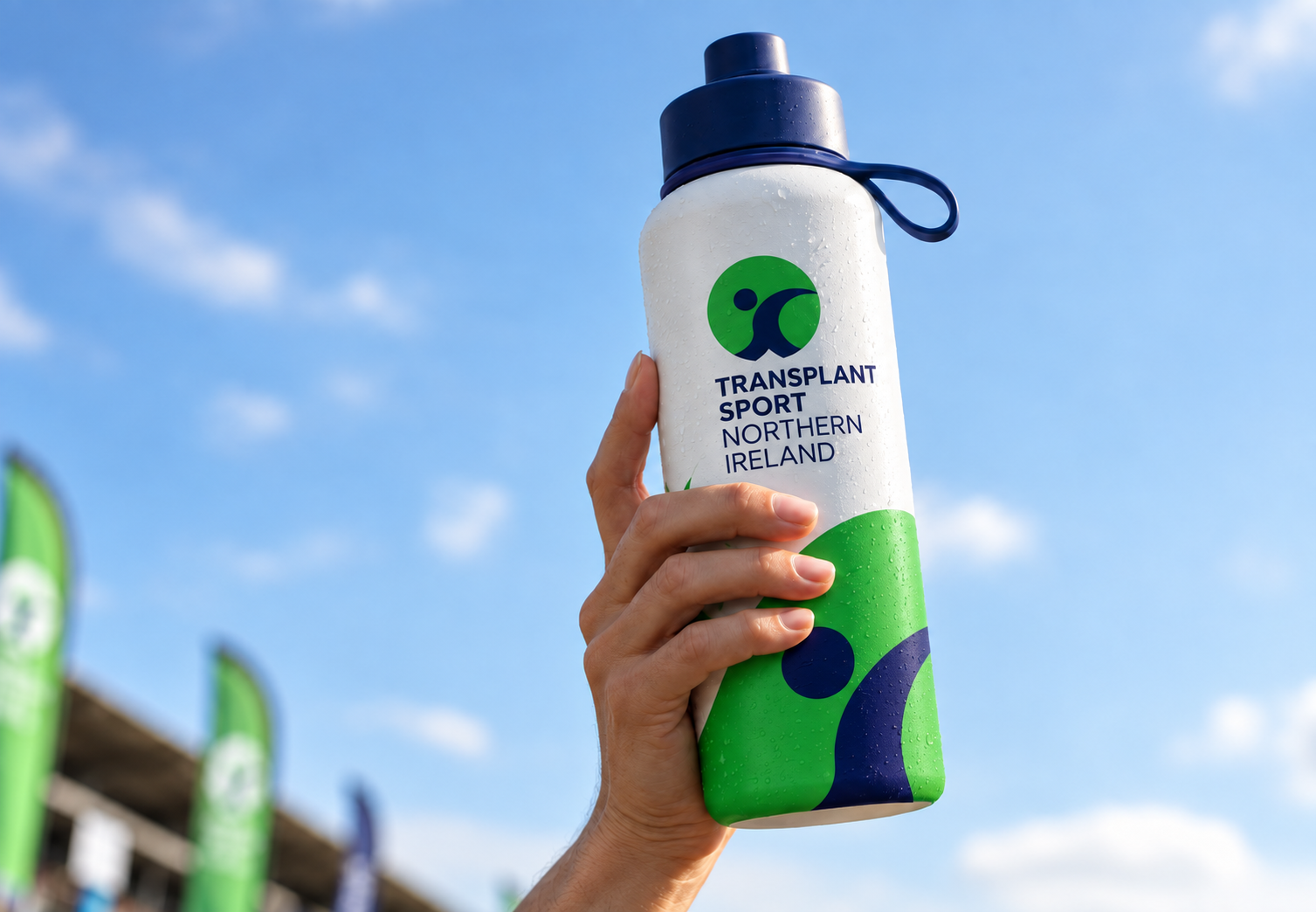 A hand holding a water bottle with the logo of Transplant Sport Northern Ireland against a blue sky with a few clouds, and green banners visible in the background.