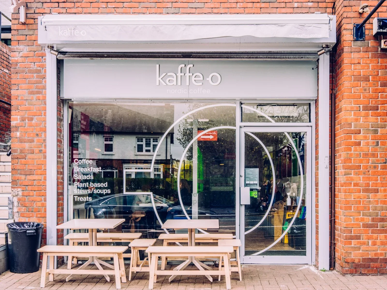 Front view of a Nordic coffee shop with a glass window and door, outdoor seating with three wooden tables and benches, and a brick exterior. The shop's signage says "kaffe.o" and lists offerings such as coffee, breakfast, salads, plant-based stews, soups, and treats.