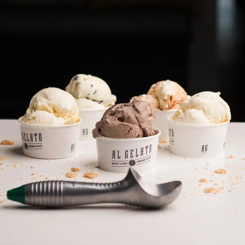 Five cups of different flavors of gelato with a small ice cream scoop in front, against a dark background.