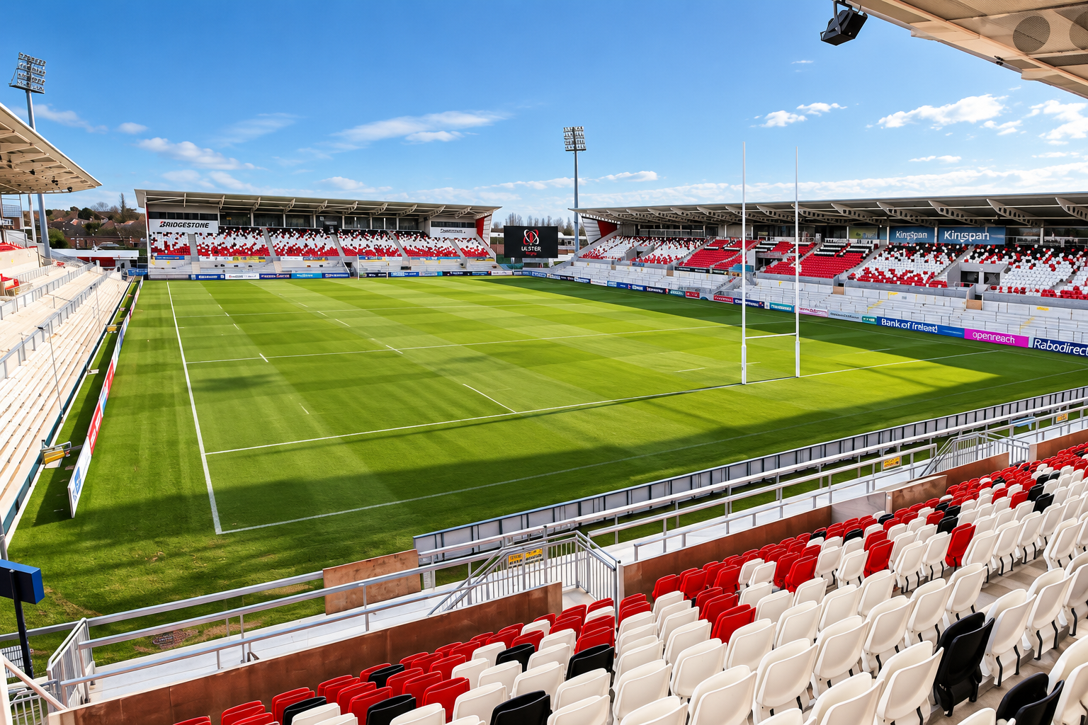 Empty rugby stadium with green field, white lines, goal posts, stands with red, white, and black seats, and blue sky.