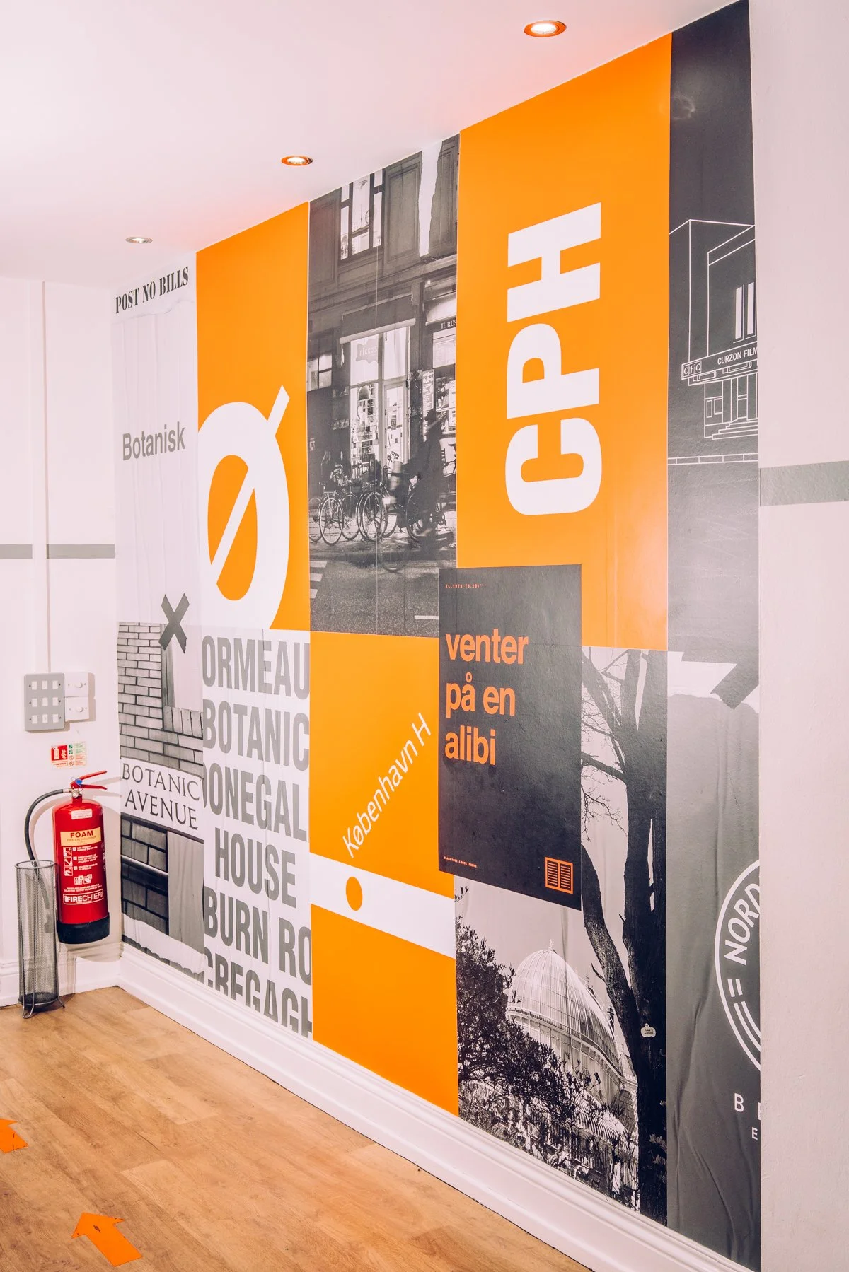 Colorful wall mural with black and white cityscape photo, large orange and black text reading 'CPH' and 'venter på en albi', and various words and images related to Copenhagen, including the Copenhagen Botanical Garden, with a fire extinguisher and fire alarm on the wall, and orange arrows pointing towards the door.
