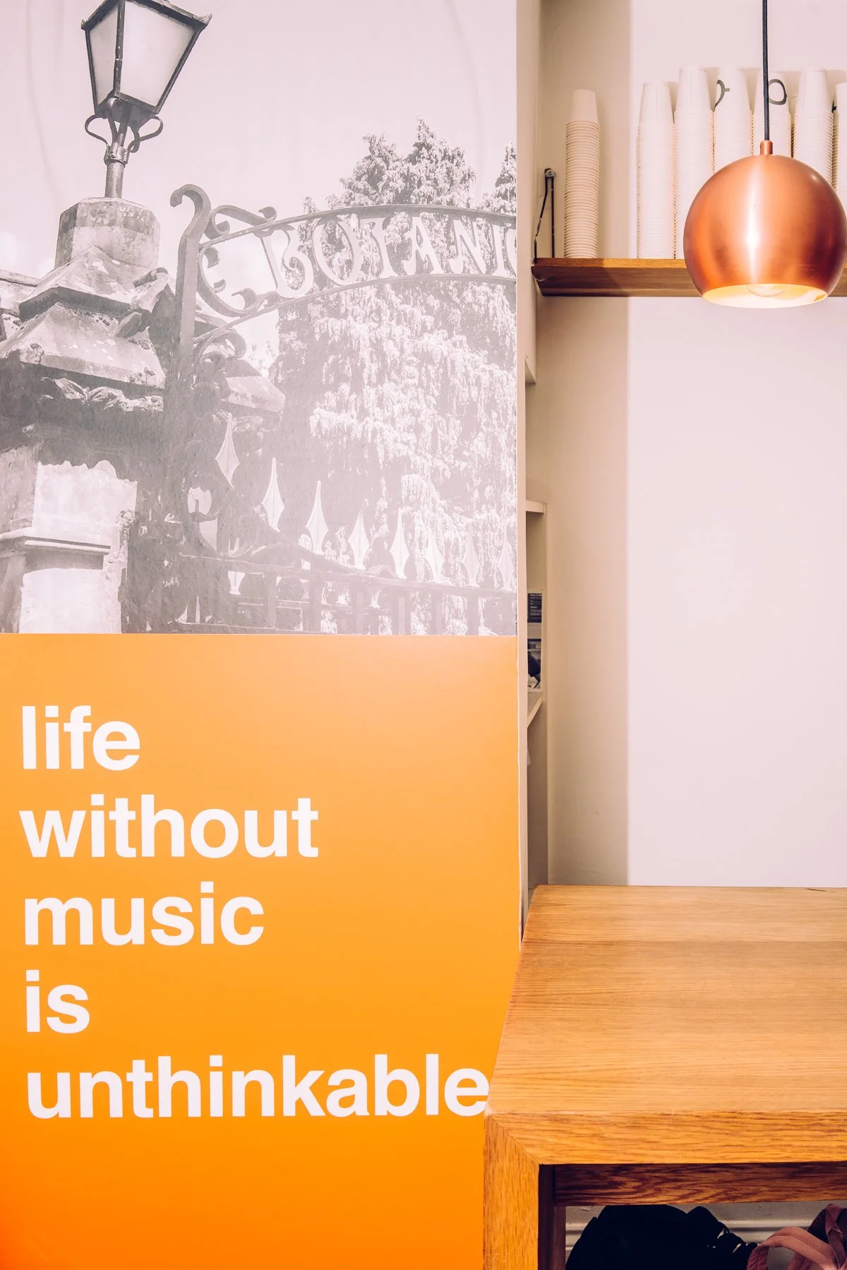Interior of a cafe with a large poster that reads 'life without music is unthinkable' and a wooden table. The poster features a black and white image of a street lamp and an ornate gate. A copper-colored pendant light hangs above the table. In the background, shelves with stacked disposable cups are visible.