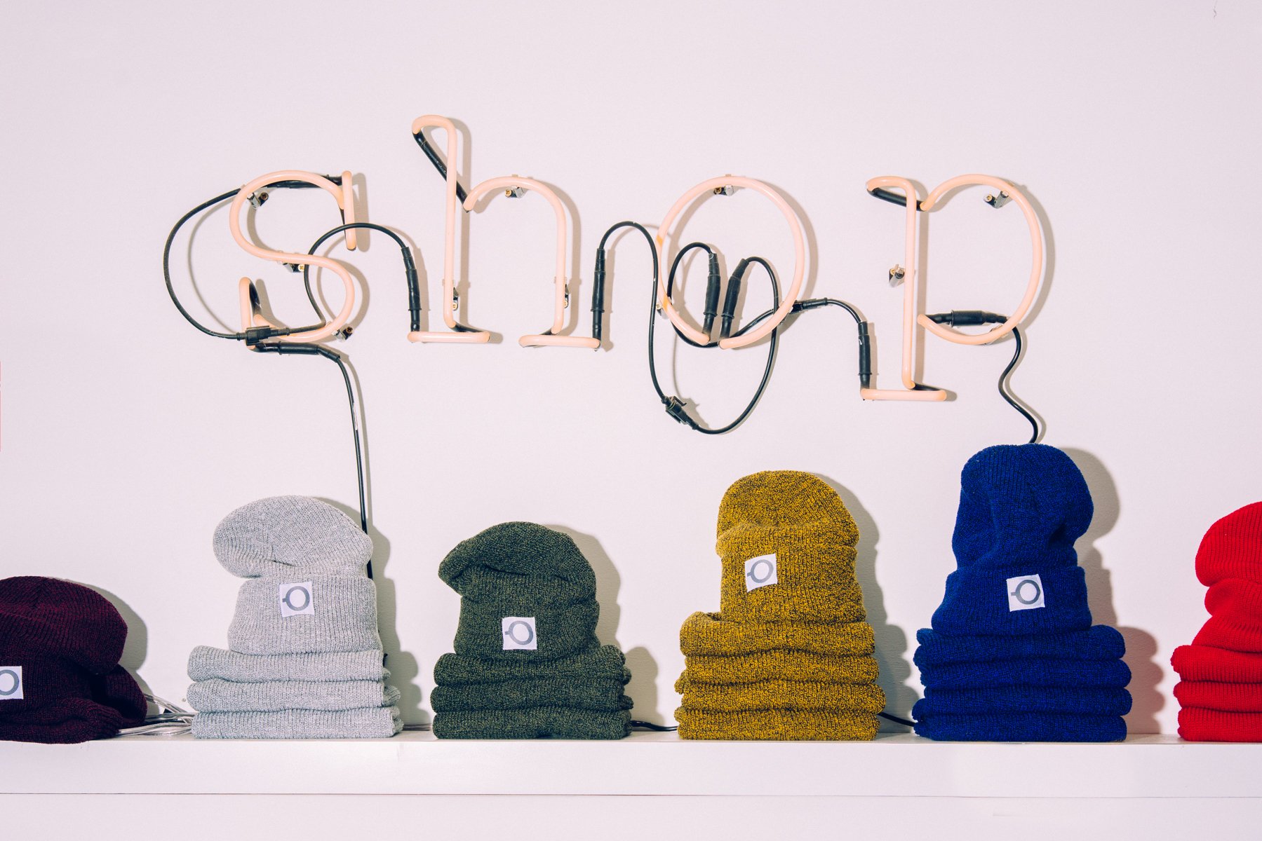 Colorful bundled beanies stacked on a white surface with a white wall and neon sign spelling 'style' in the background.
