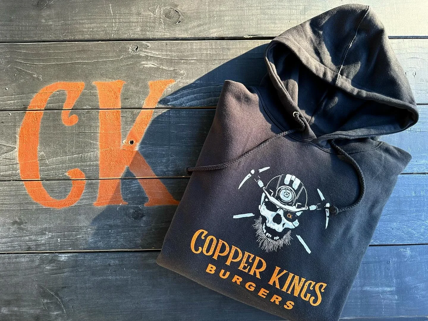 A black hoodie with a skeleton wearing a miner's helmet and the text 'Copper Kings Burgers' printed on it, lying on a wooden surface with the words 'CK' and 'K' painted in orange.