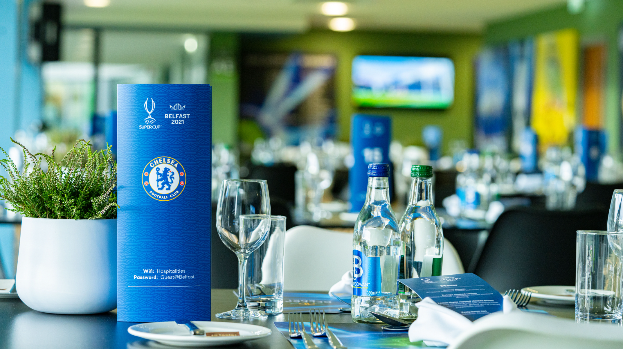 A dinner table at a restaurant decorated with Chelsea Football Club promotional materials, including a blue menu and bottles of water, with a potted plant and wine glass in the foreground and a television screen in the background.