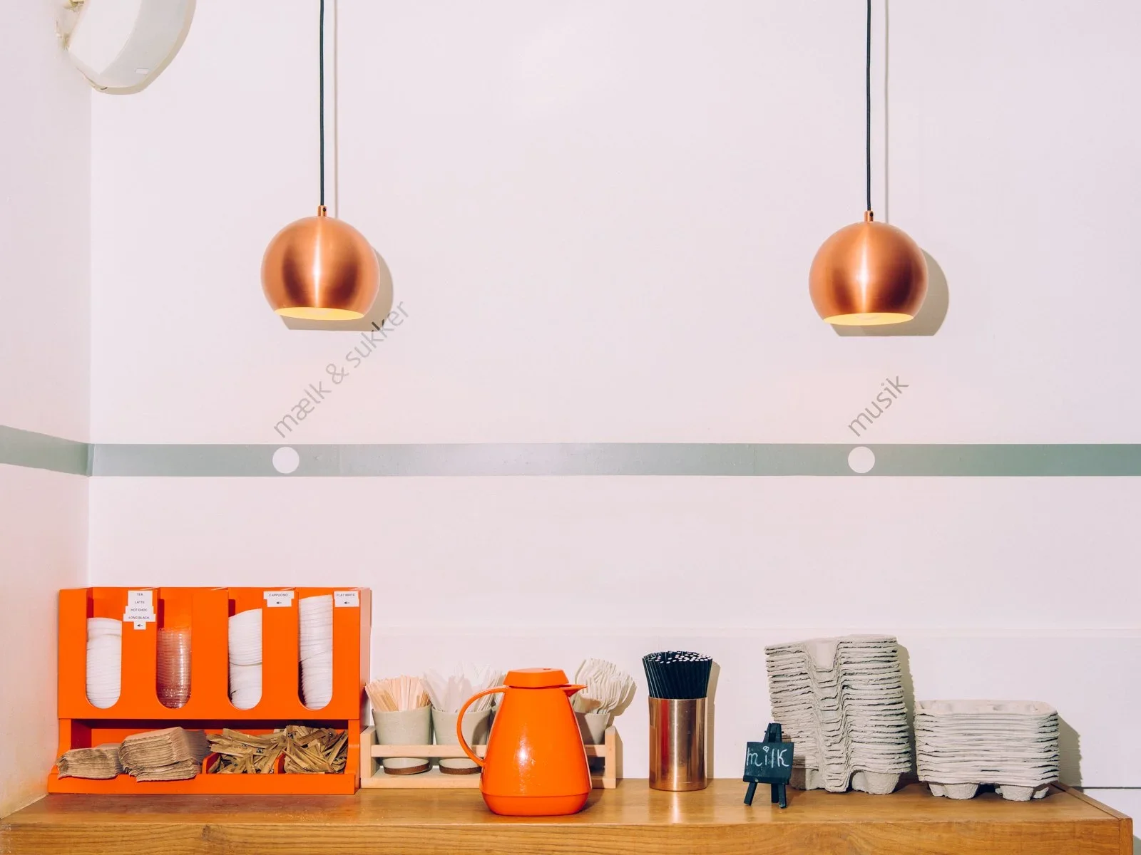 A cafe counter with stacks of napkins, disposable cups, an orange beverage dispenser, black straws in a metal container, and orange and white boxes for tea or coffee. Two copper pendant lights hang from the ceiling, illuminating the area.