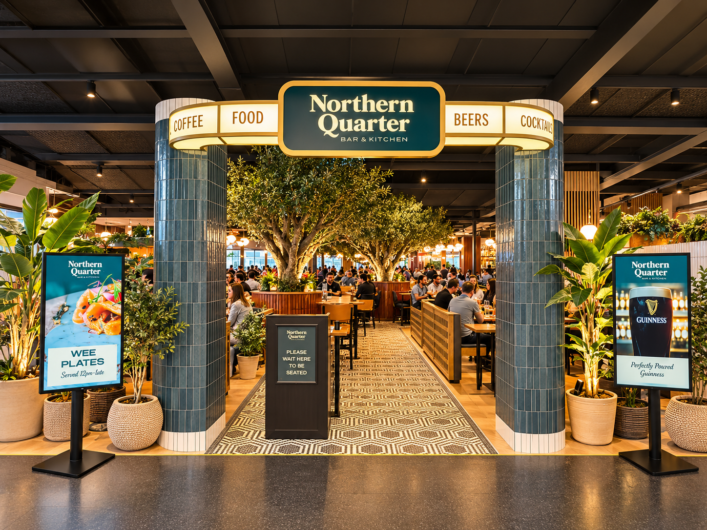 Entrance to Northern Quarter bar and kitchen with indoor seating, large trees, plants, digital menu boards, and signs advertising food and drinks.