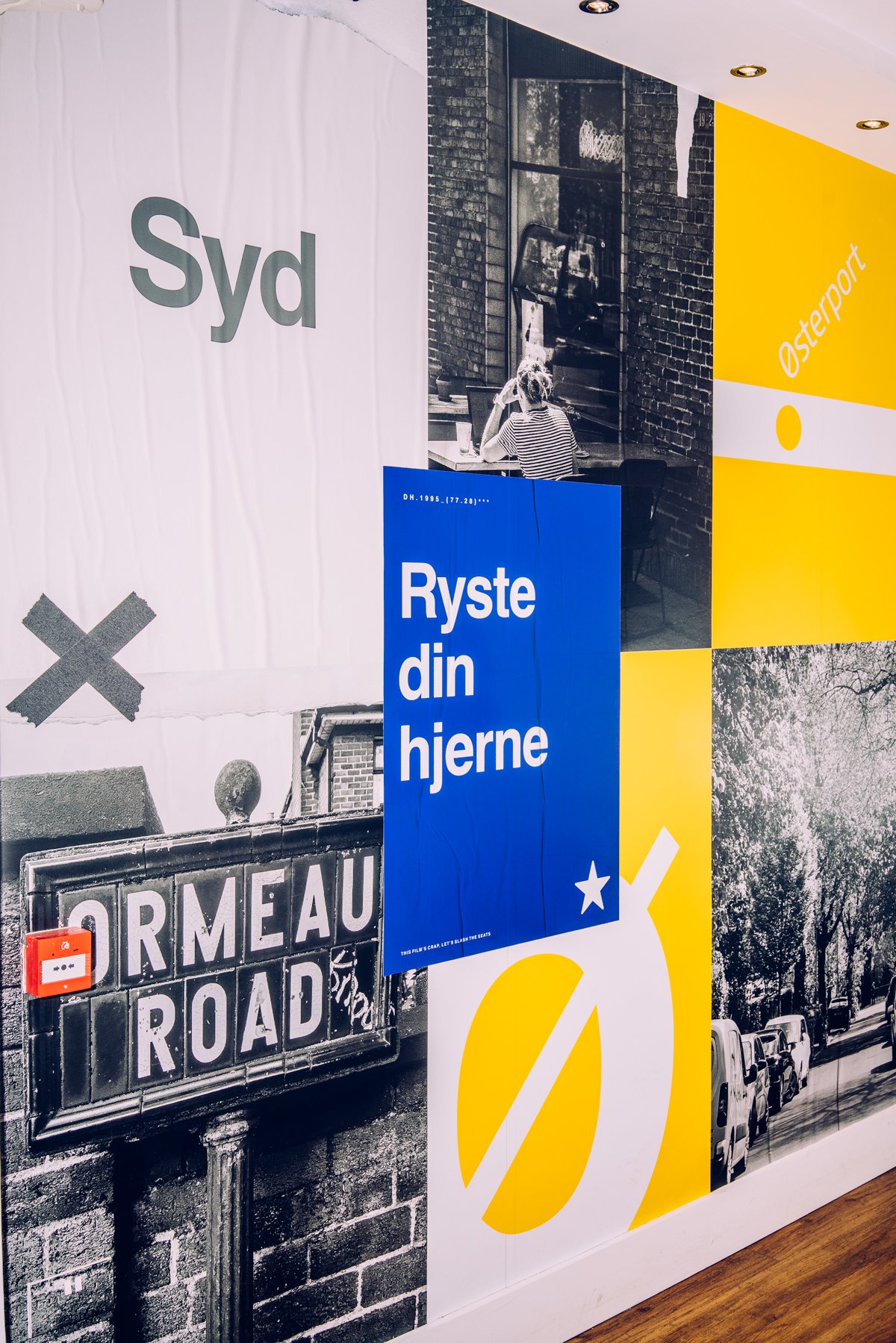 Combination of black-and-white and colored posters on a wall, including street signs, a photograph of a woman sitting in a cafe, and various graphic designs with text in Danish and English.