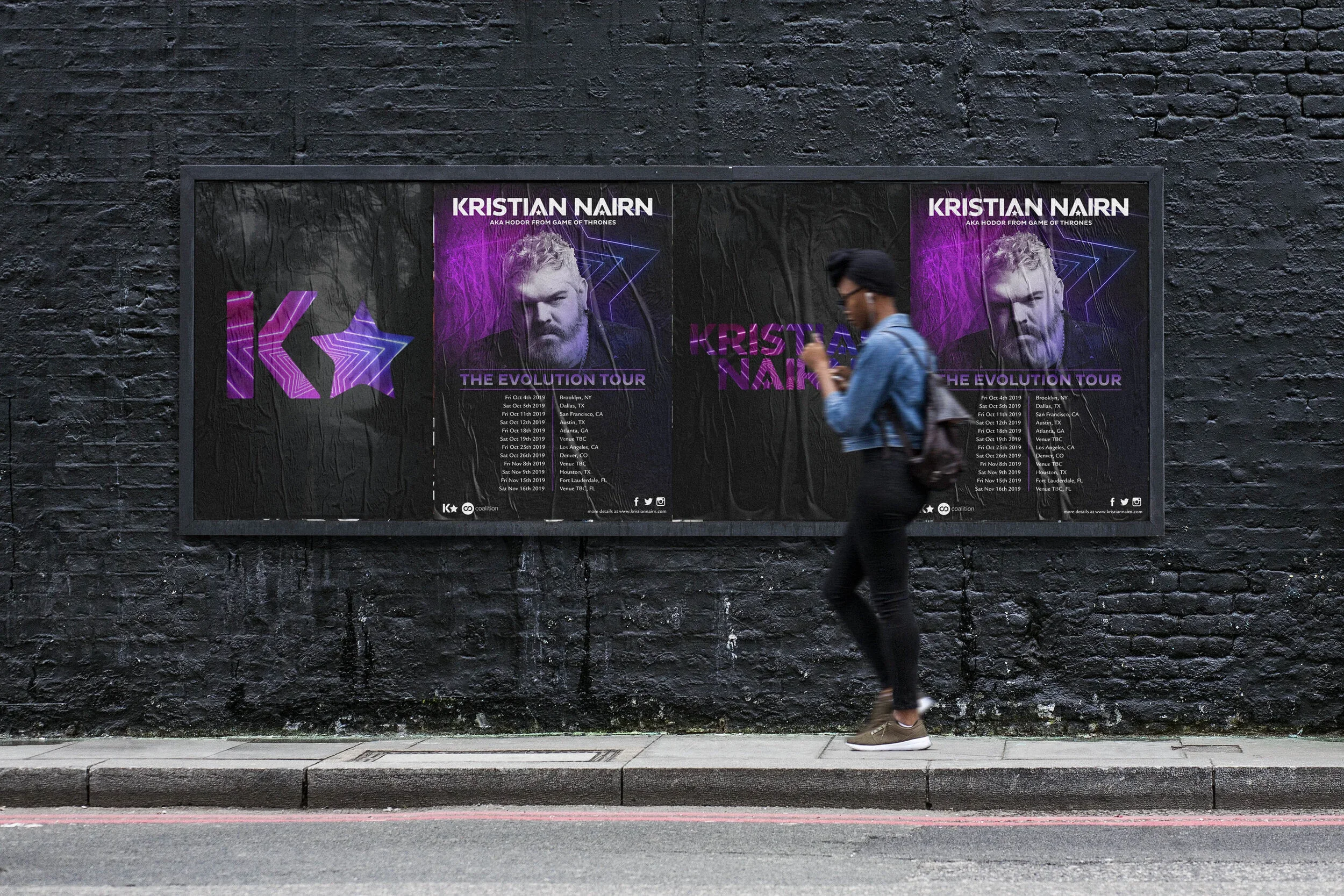 A man walking on a city sidewalk past a large poster on a black brick wall. The poster promotes Kristian Nairn's 'The Evolution Tour,' featuring his image, tour dates, and locations.